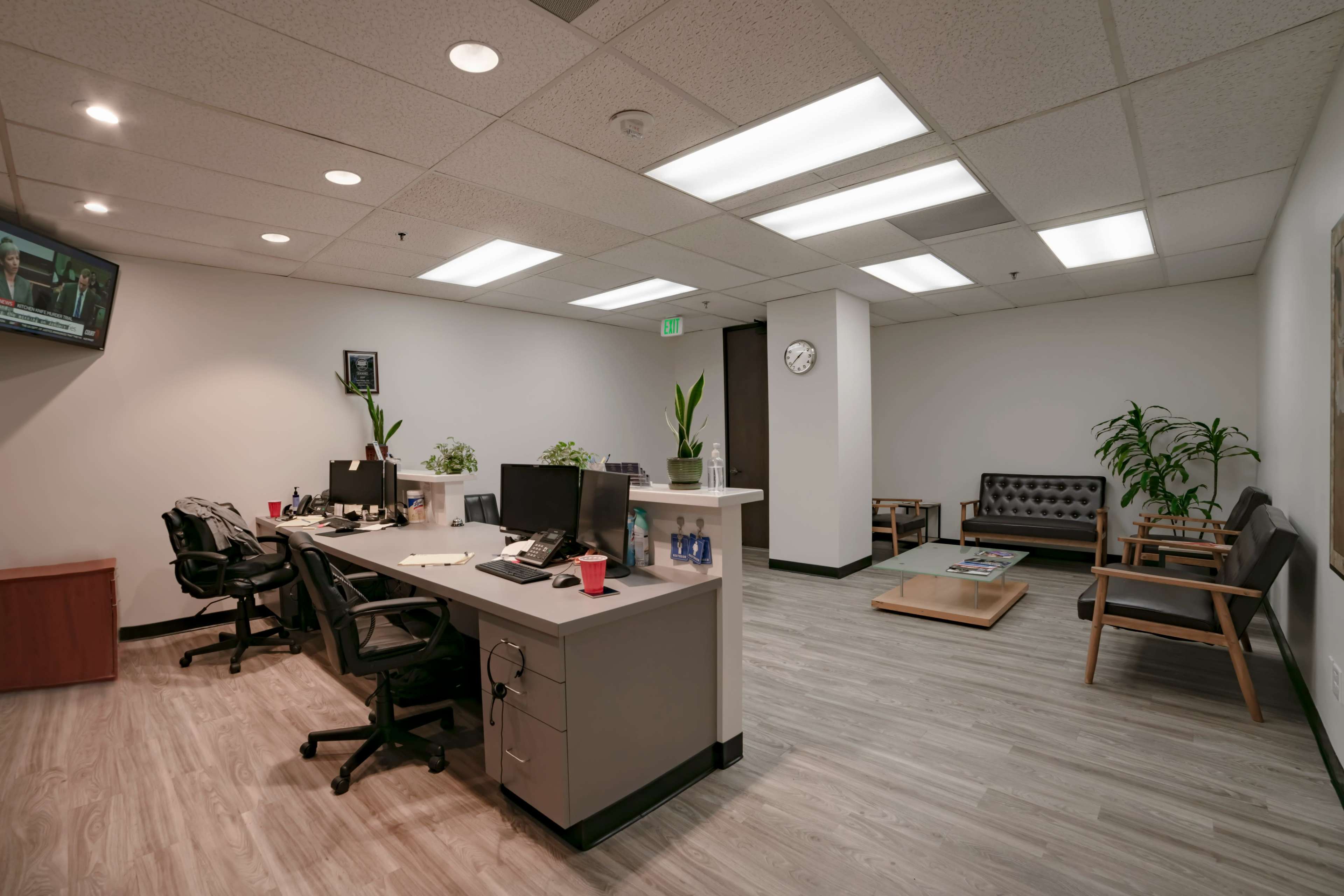A modern office space with two workstations, a waiting area with chairs, and television displaying news on the wall.