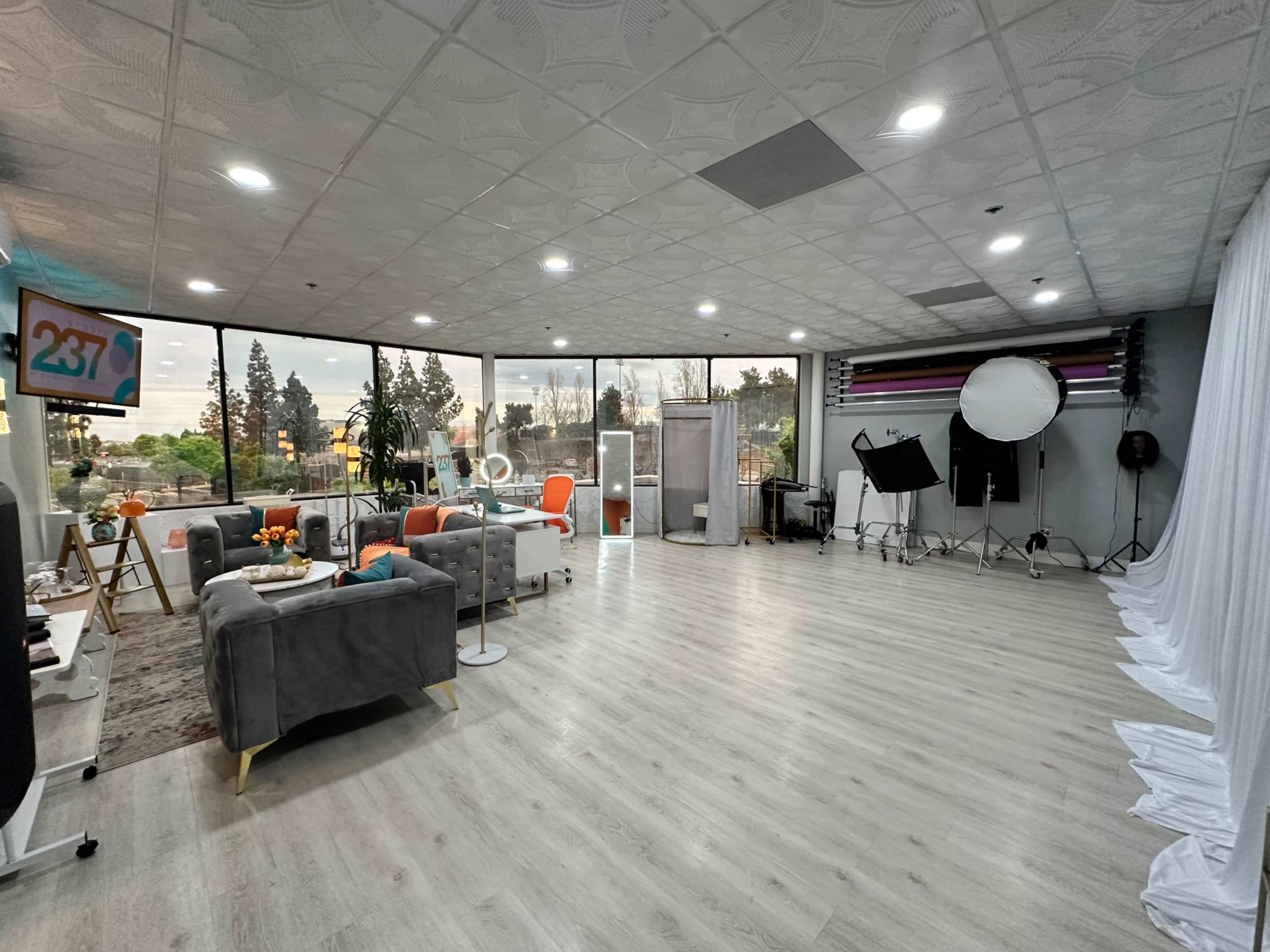 The image shows a spacious, well-lit studio with large windows, featuring a seating area, decorative plants, and photography equipment in the background.