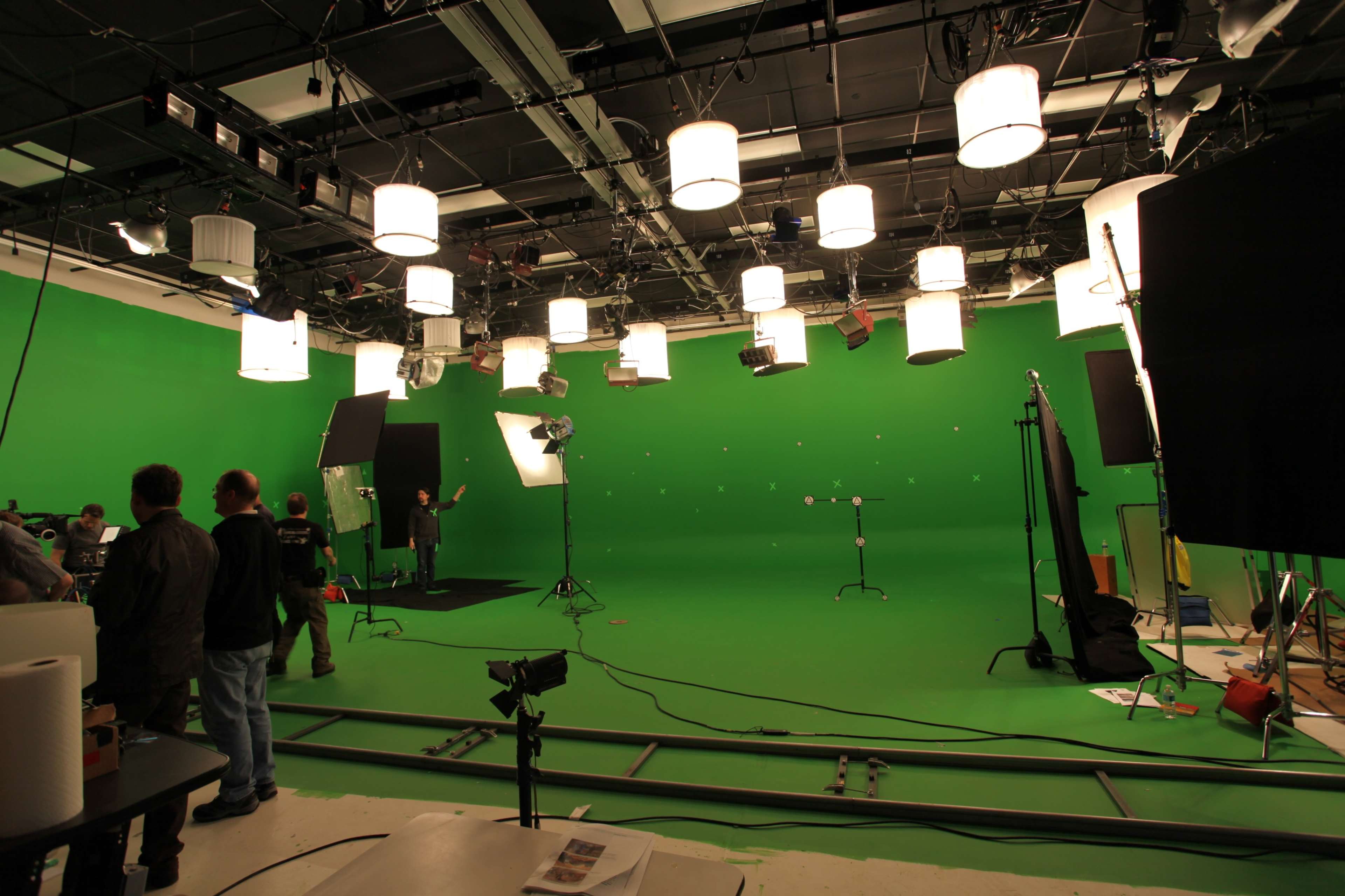 A film production set features a green screen backdrop with multiple lighting fixtures and crew members preparing for a shoot.