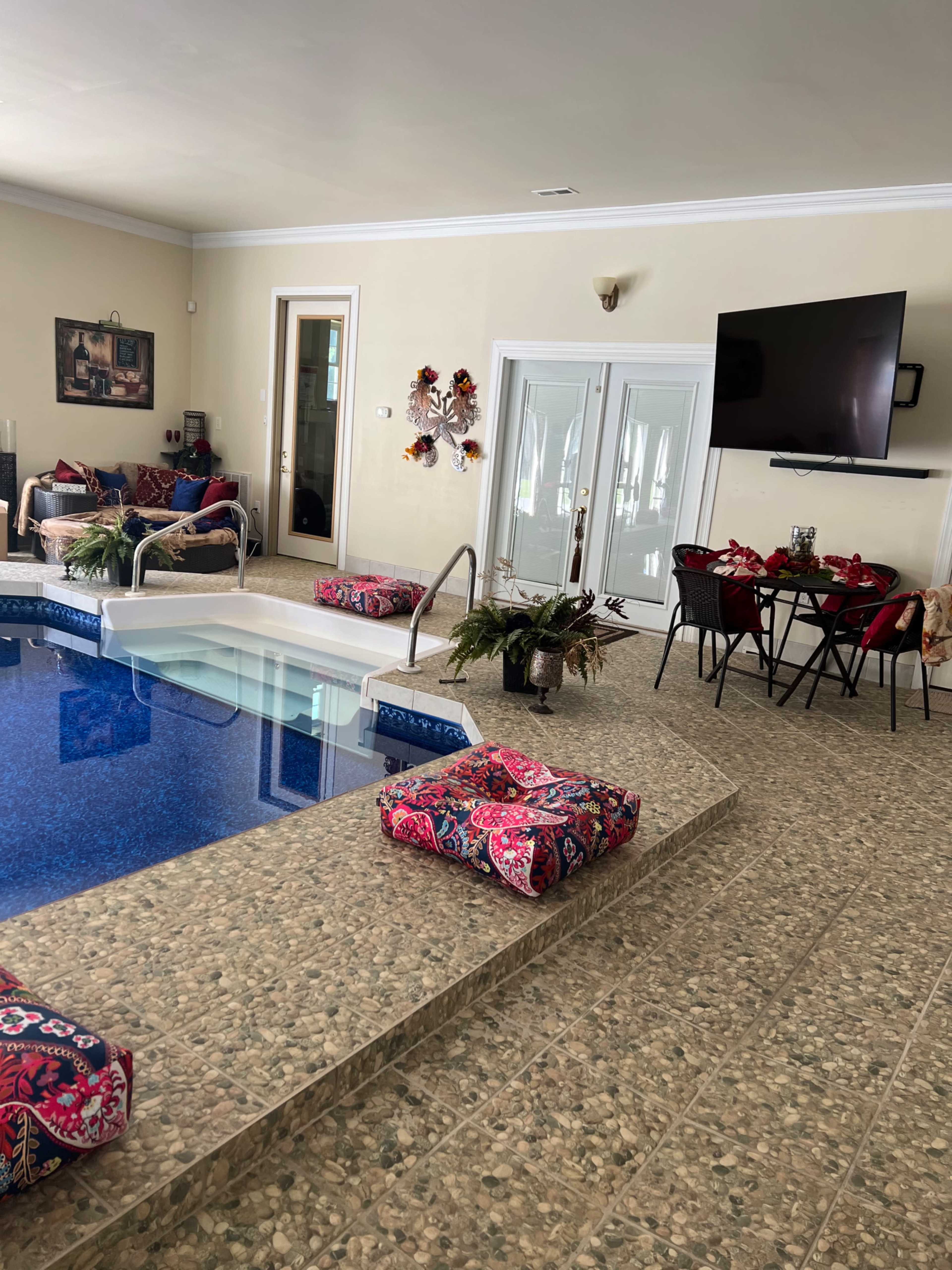 An indoor swimming pool area with lounge seating, a dining table, and a large television mounted on the wall.