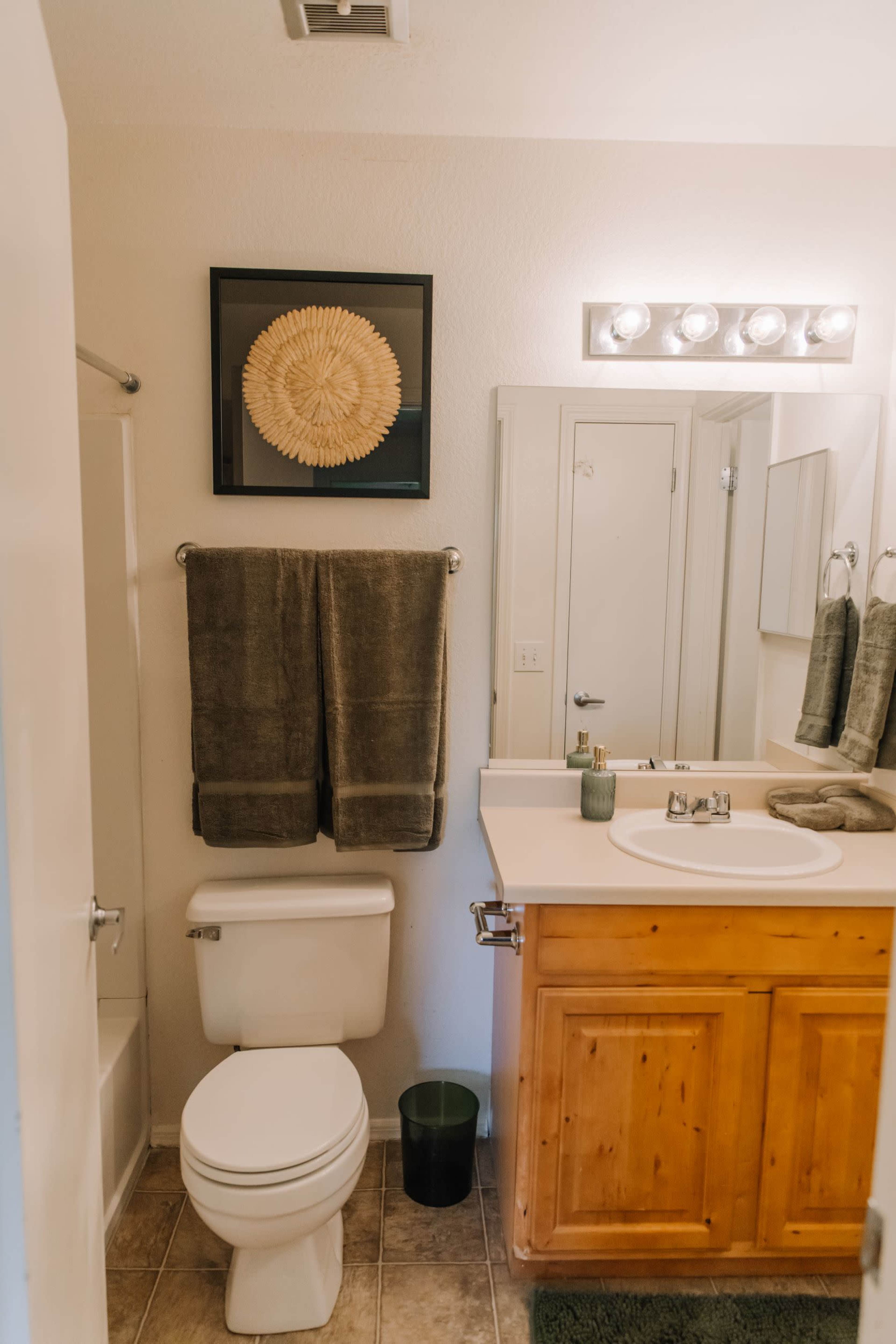 A clean bathroom features a wooden cabinet, white toilet, and a sink with a large mirror above it, complemented by gray towels and wall decor.