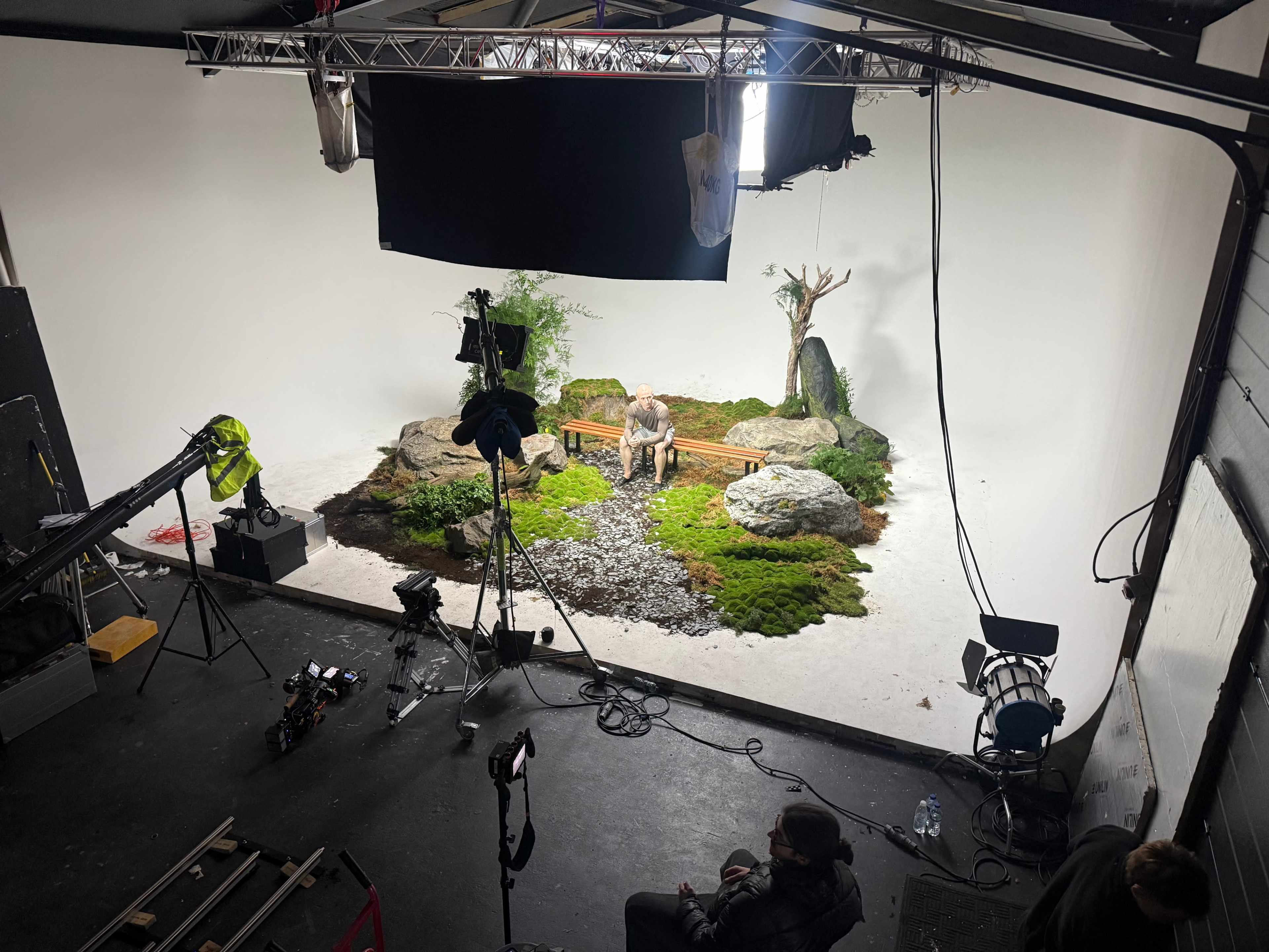 The image shows a film set with a moss-covered ground, rocks, and a bench, featuring equipment like cameras and lighting arranged around the scene.