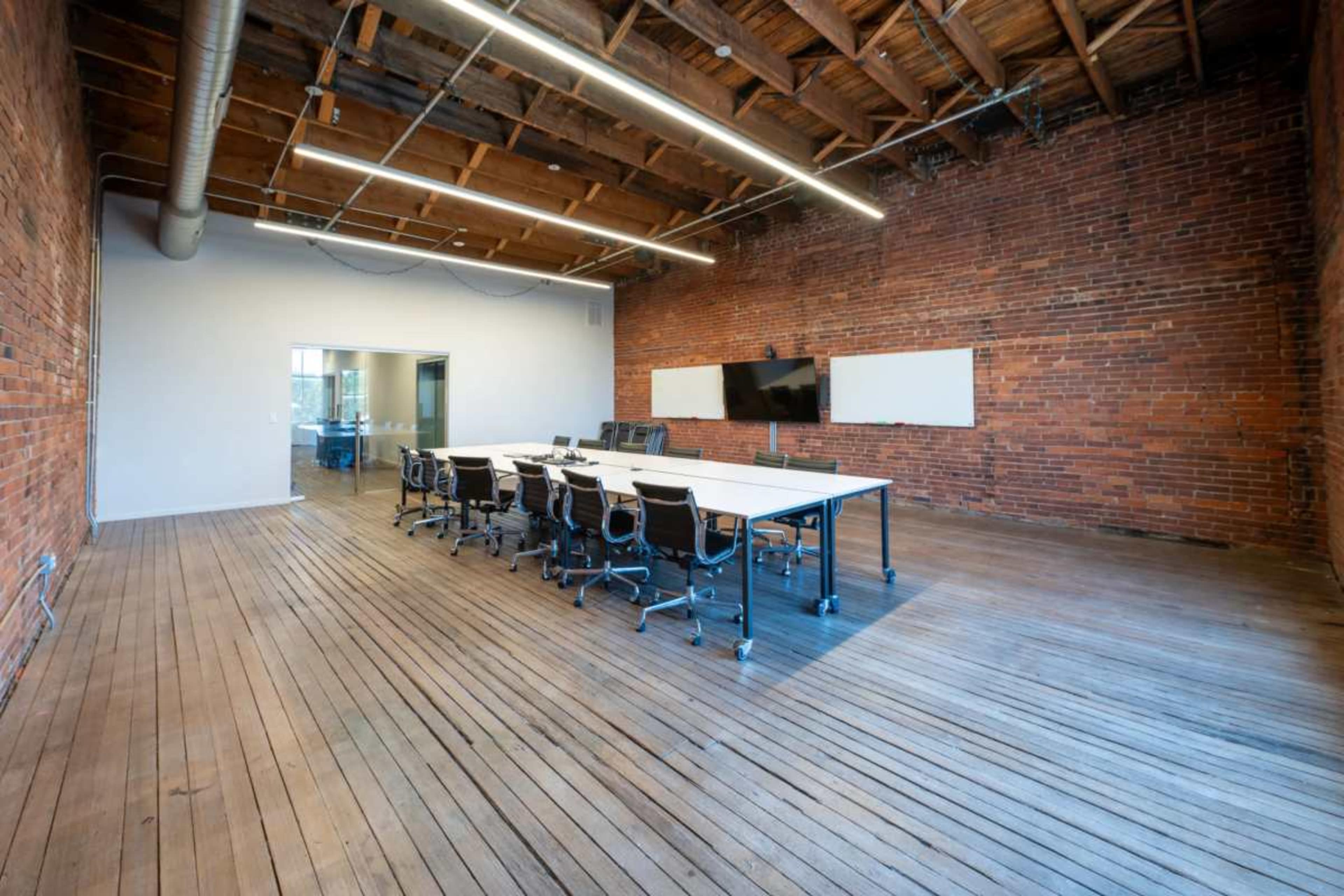 A large conference room features a long table surrounded by black chairs, with exposed brick walls and wooden flooring, along with two presentation screens.