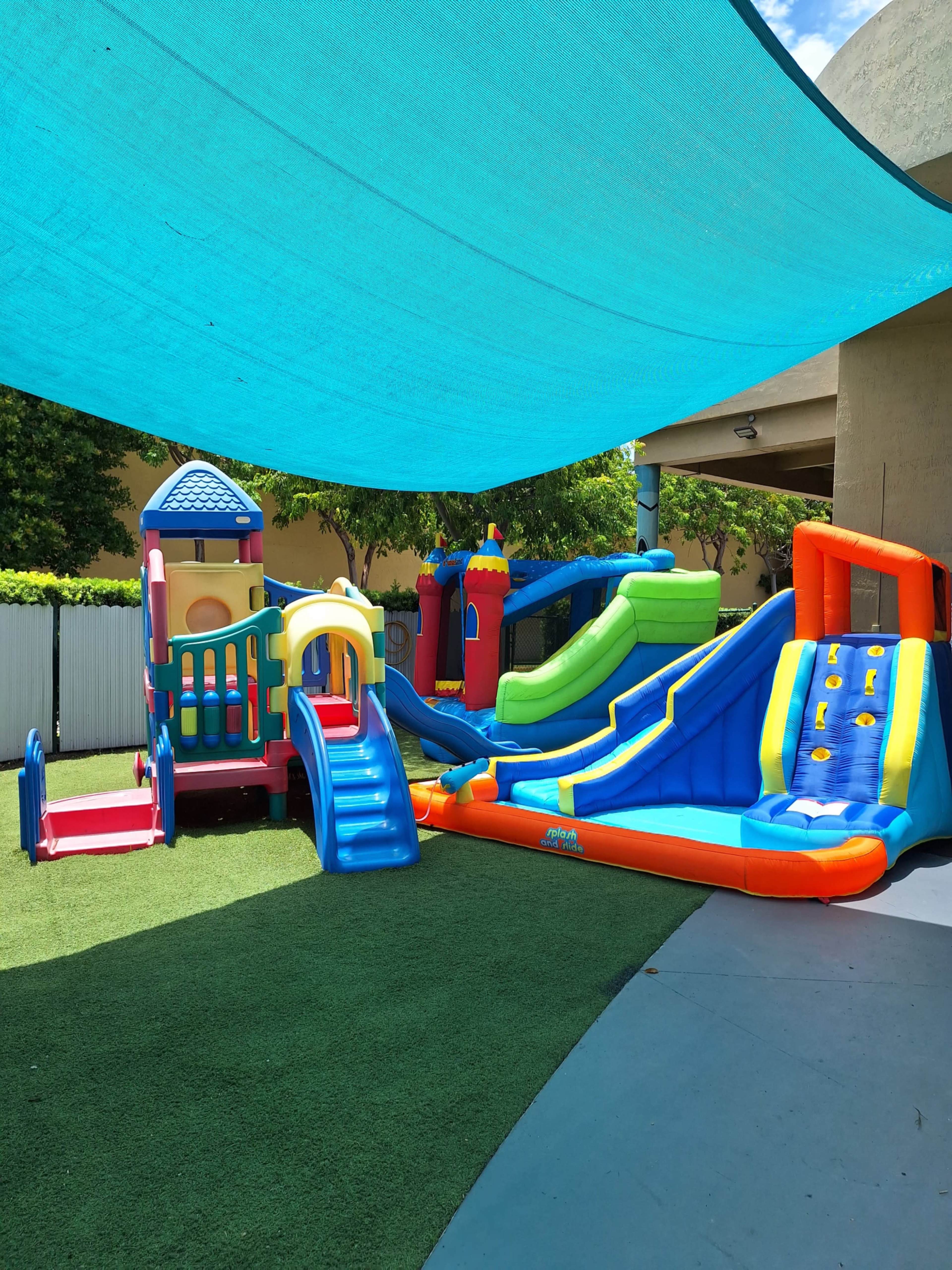 The image shows a colorful inflatable playground area with slides and climbing structures, shaded by a large blue canopy.