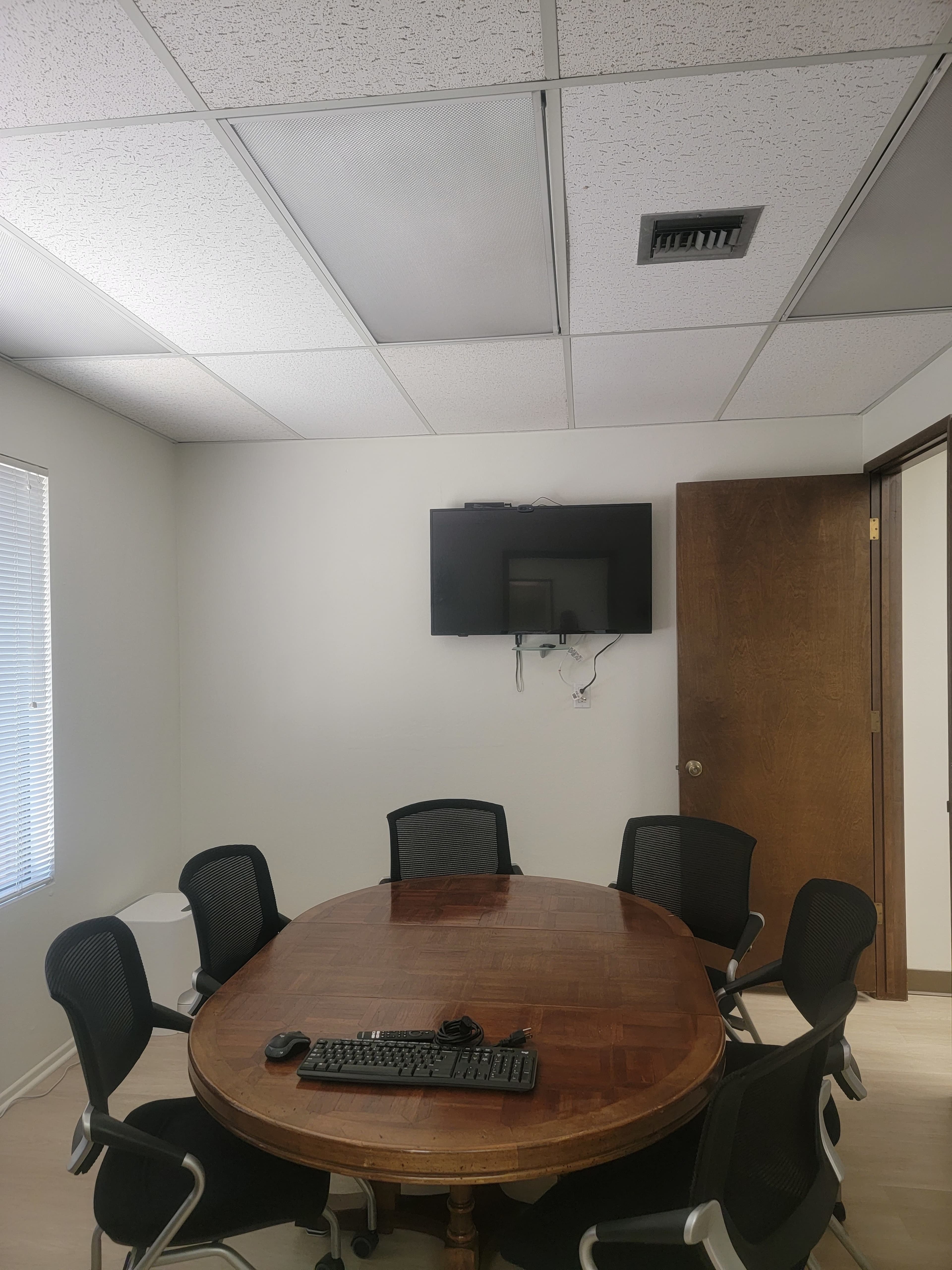 A conference room features a round wooden table surrounded by six black chairs, with a television mounted on the wall.