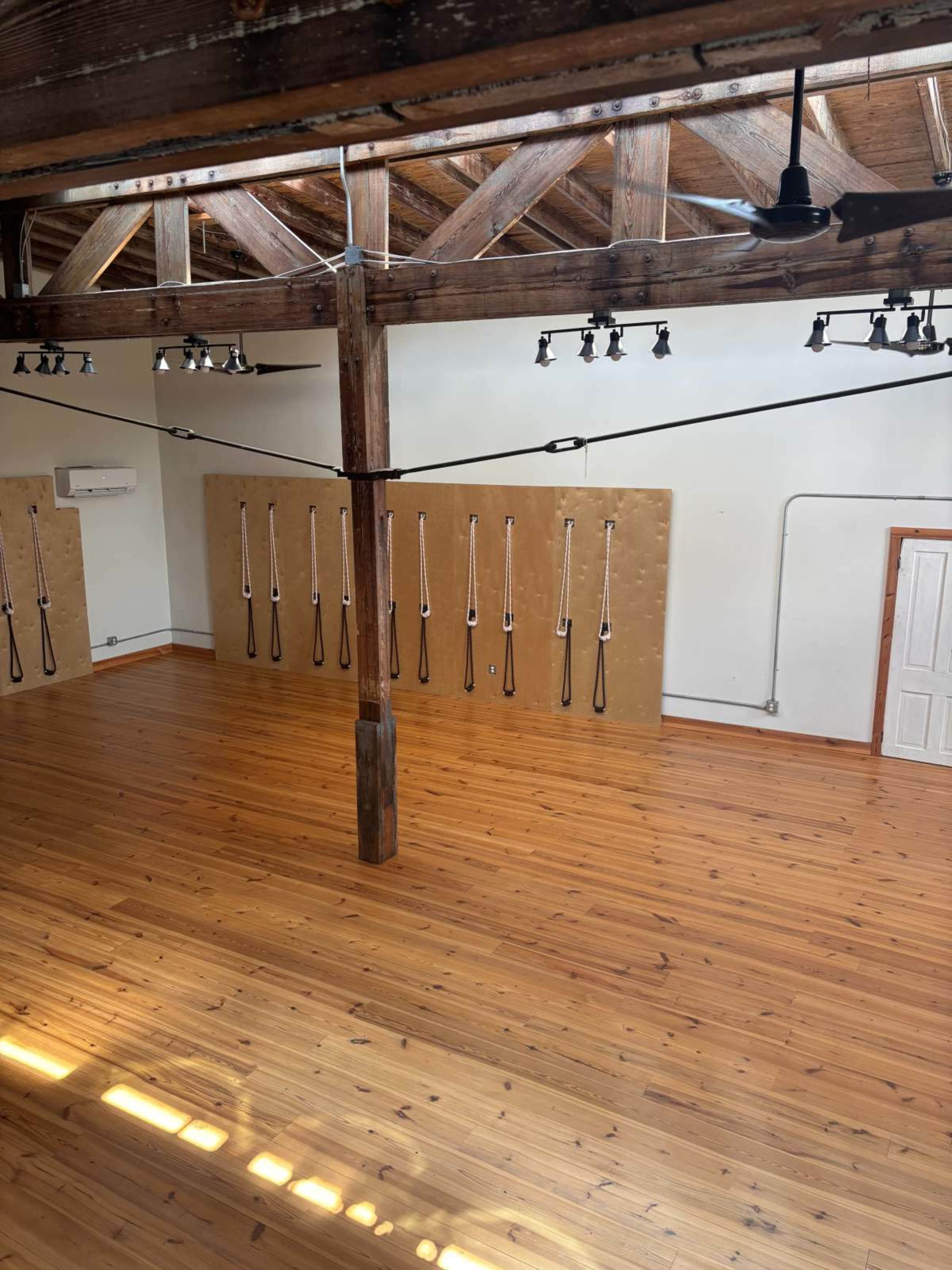 The image shows a spacious wooden-floored room with exposed beams, featuring wall-mounted exercise equipment and bright overhead lighting.