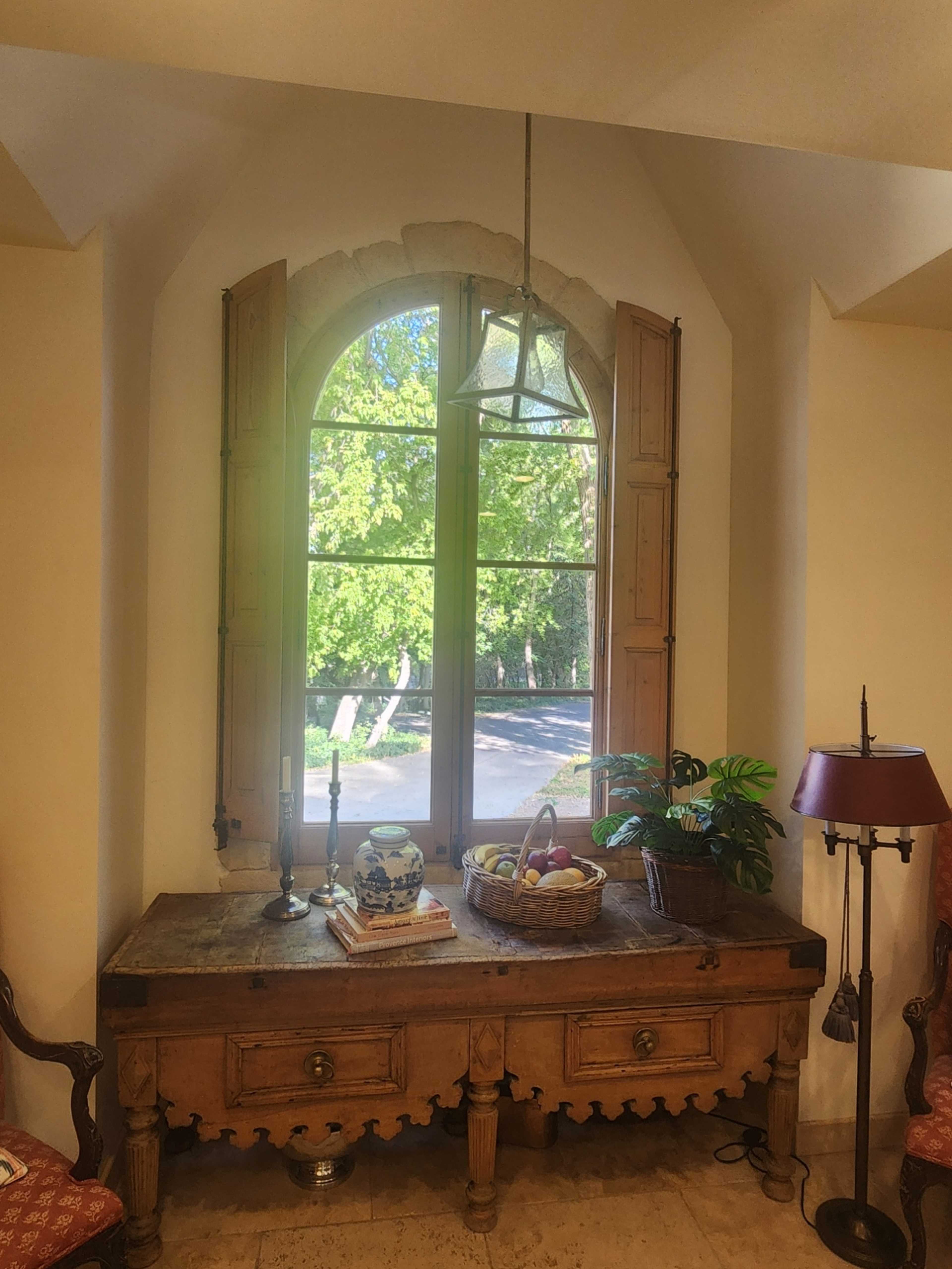 A large window with wooden shutters frames a rustic wooden table adorned with a fruit basket and decorative items, set against a backdrop of greenery outside.