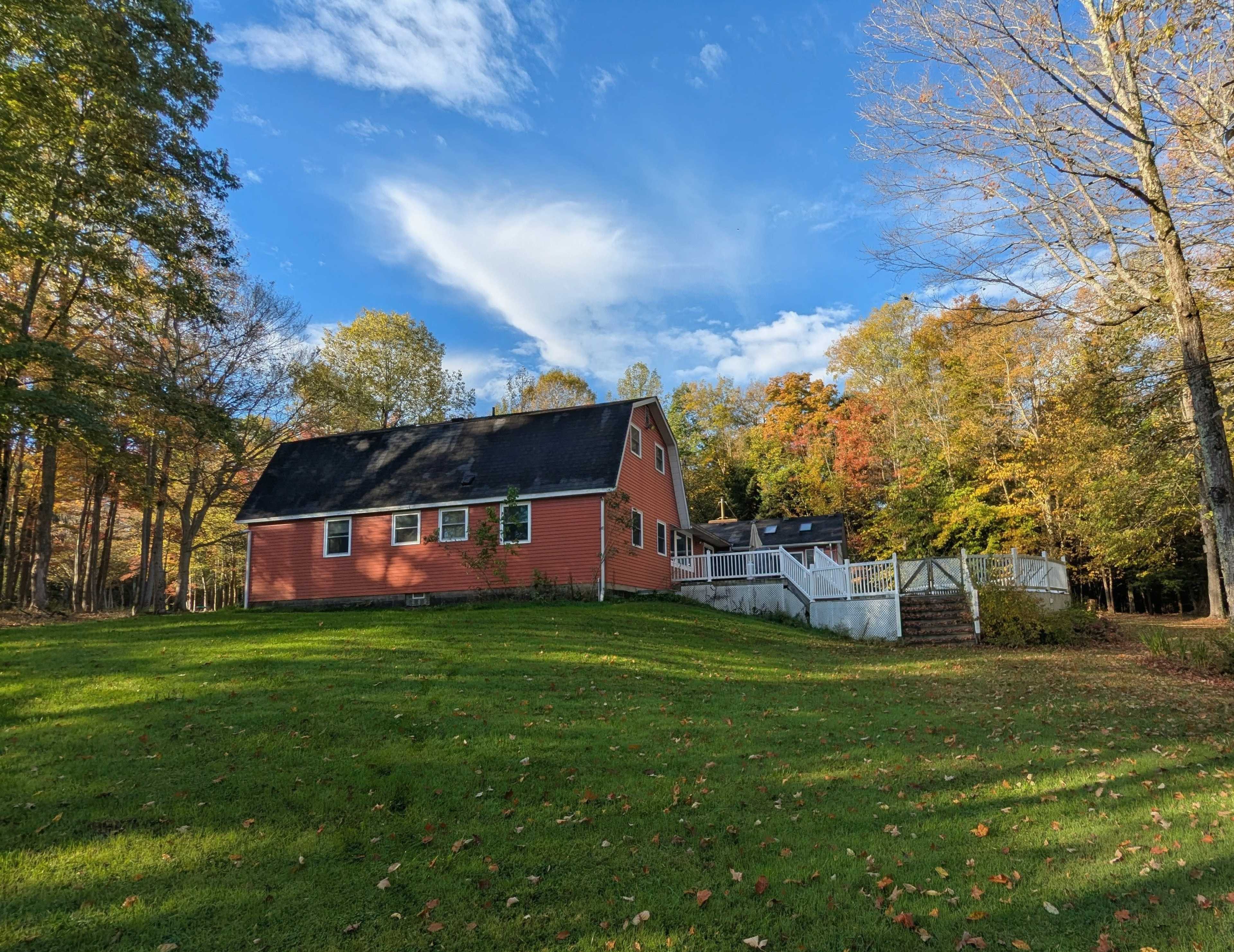 A red house with a sloped roof is situated on a grassy area surrounded by trees with autumn foliage.