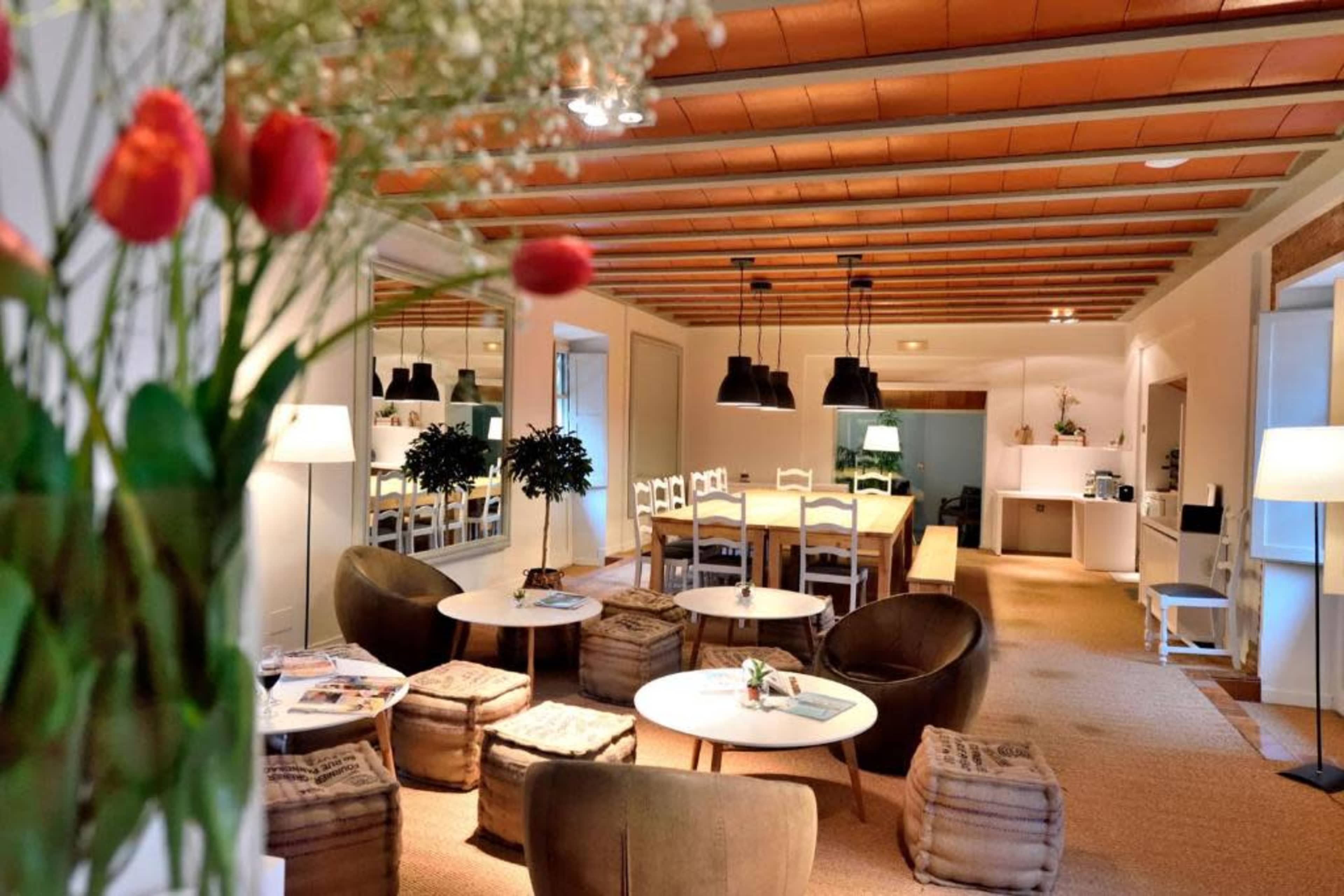 The image shows a well-lit lounge area featuring circular tables, comfortable seating, and a dining space with wooden beams and decorative plants.