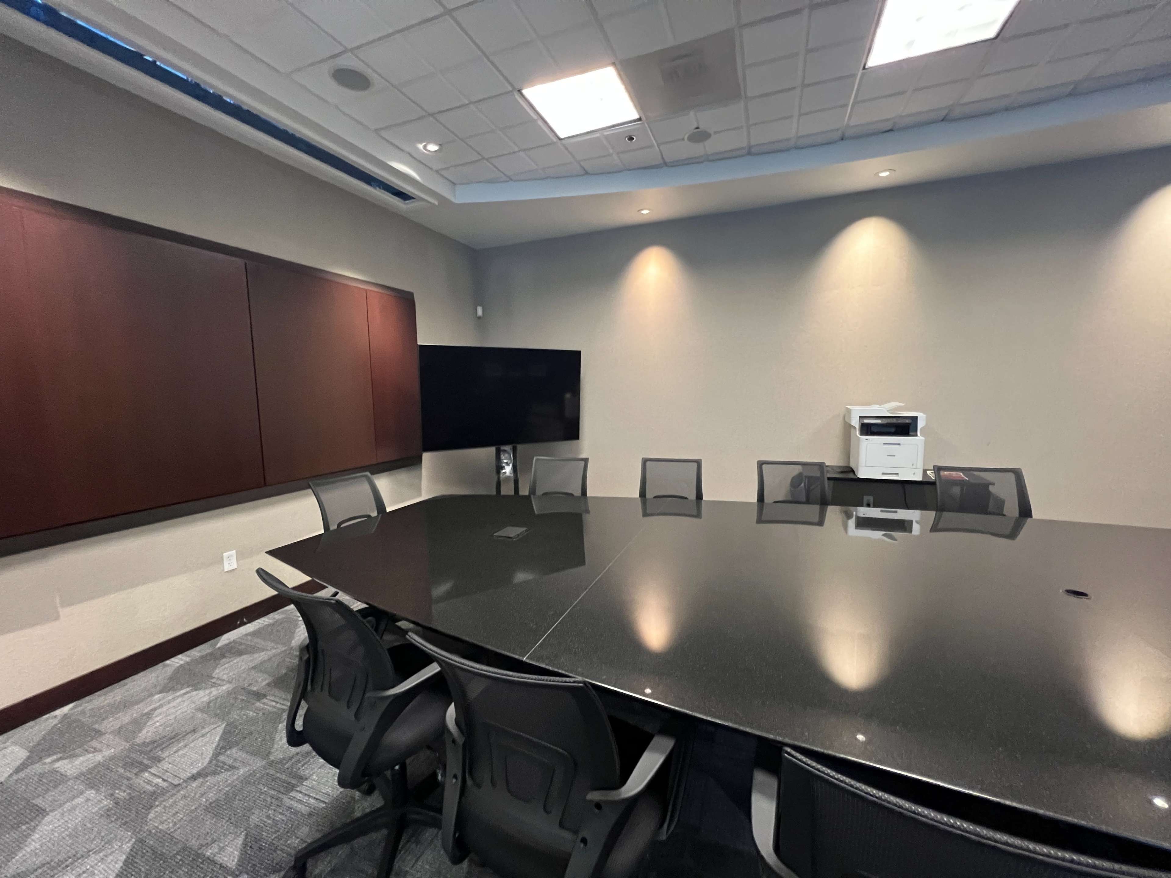 A modern conference room featuring a large black table surrounded by chairs, a wall-mounted screen, and a printer in the corner.