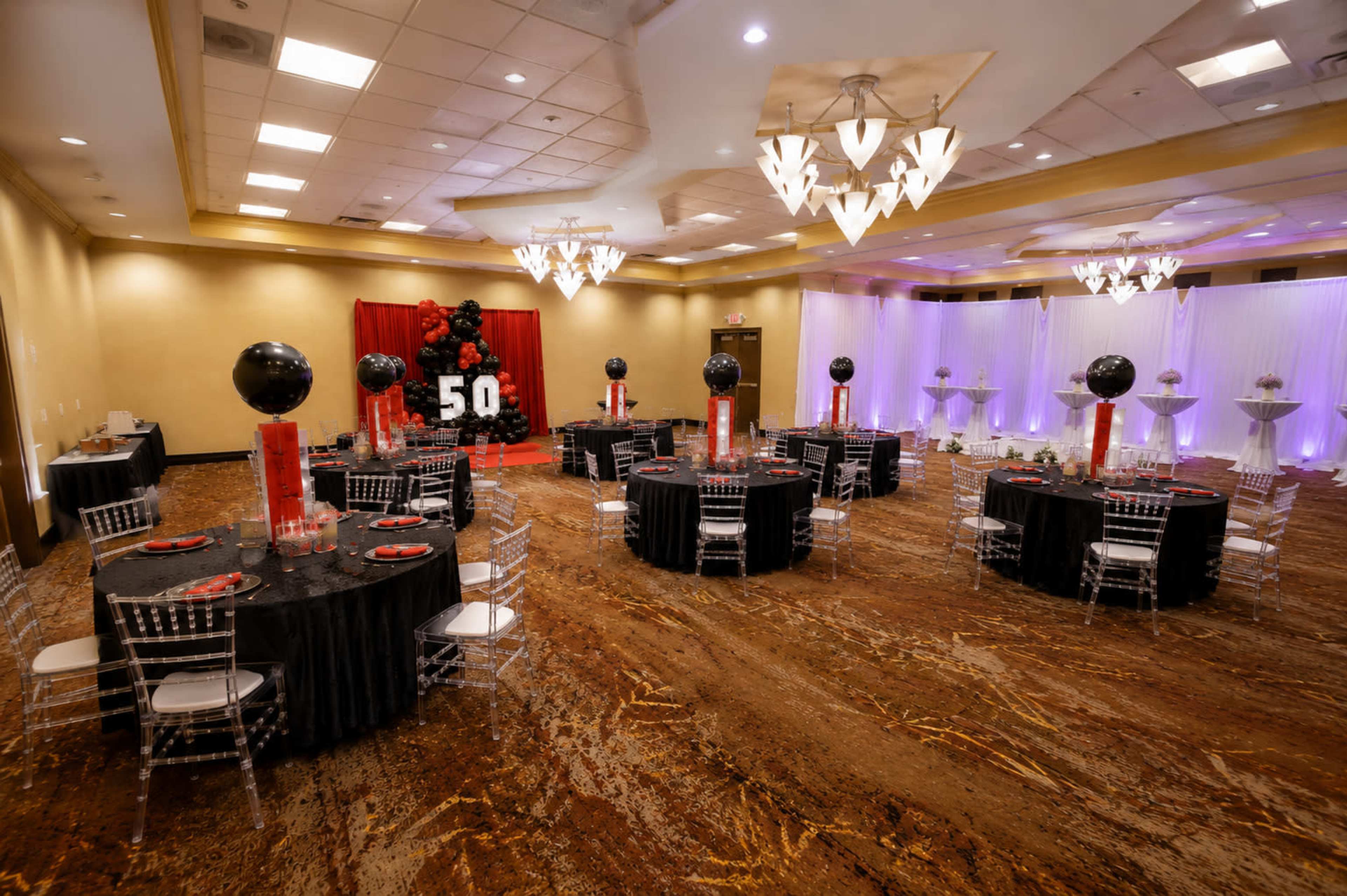 The image shows a banquet hall set up for a celebration, featuring tables dressed in black cloth, red accents, and a large decorative backdrop that reads "50."