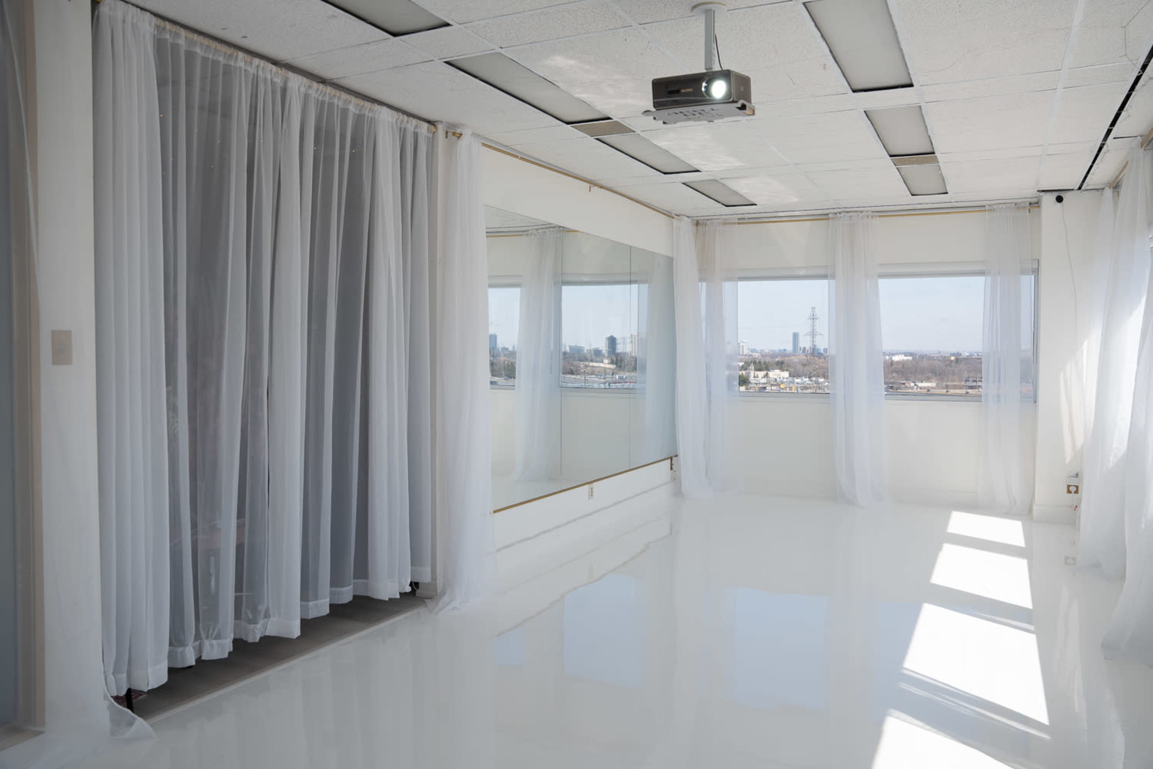 The image shows a bright, empty room with white reflective floors, sheer curtains, large mirrors, and a projector mounted on the ceiling.