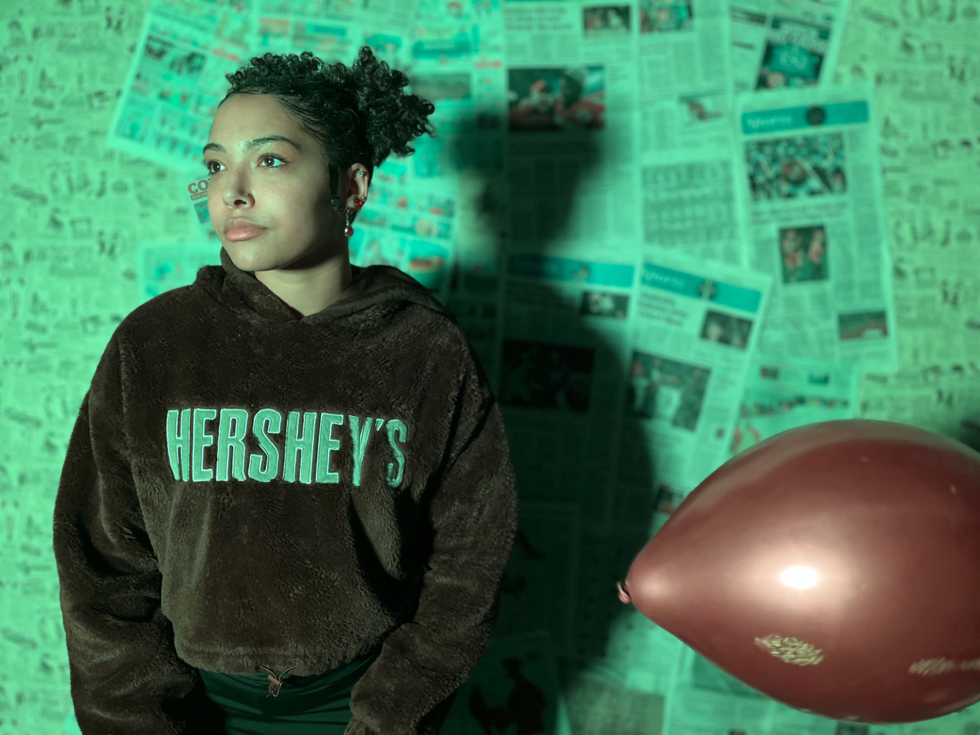 A person wearing a Hershey's hoodie stands in front of a wall covered with newspaper articles, with a red balloon nearby.
