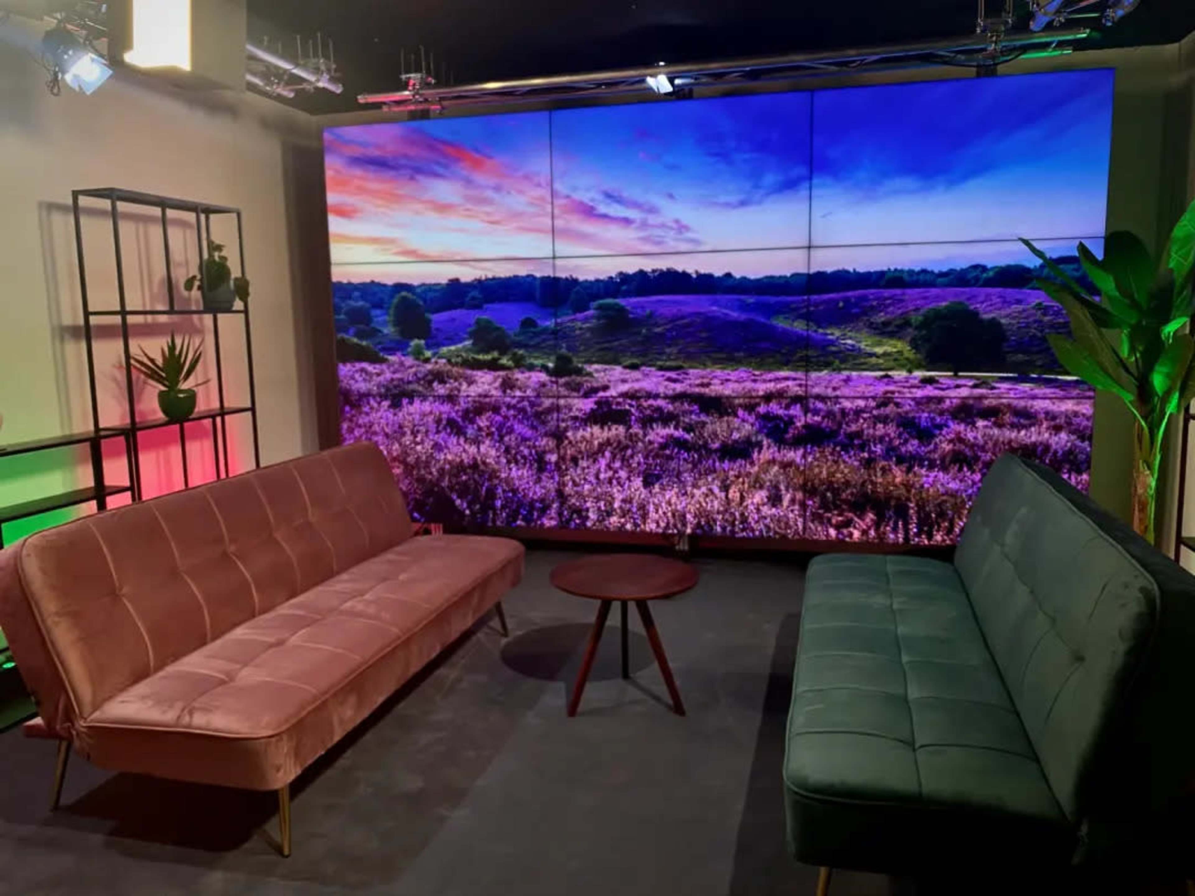 A television studio set features two couches facing a large screen displaying a scenic landscape with purple flowers and a sunset.