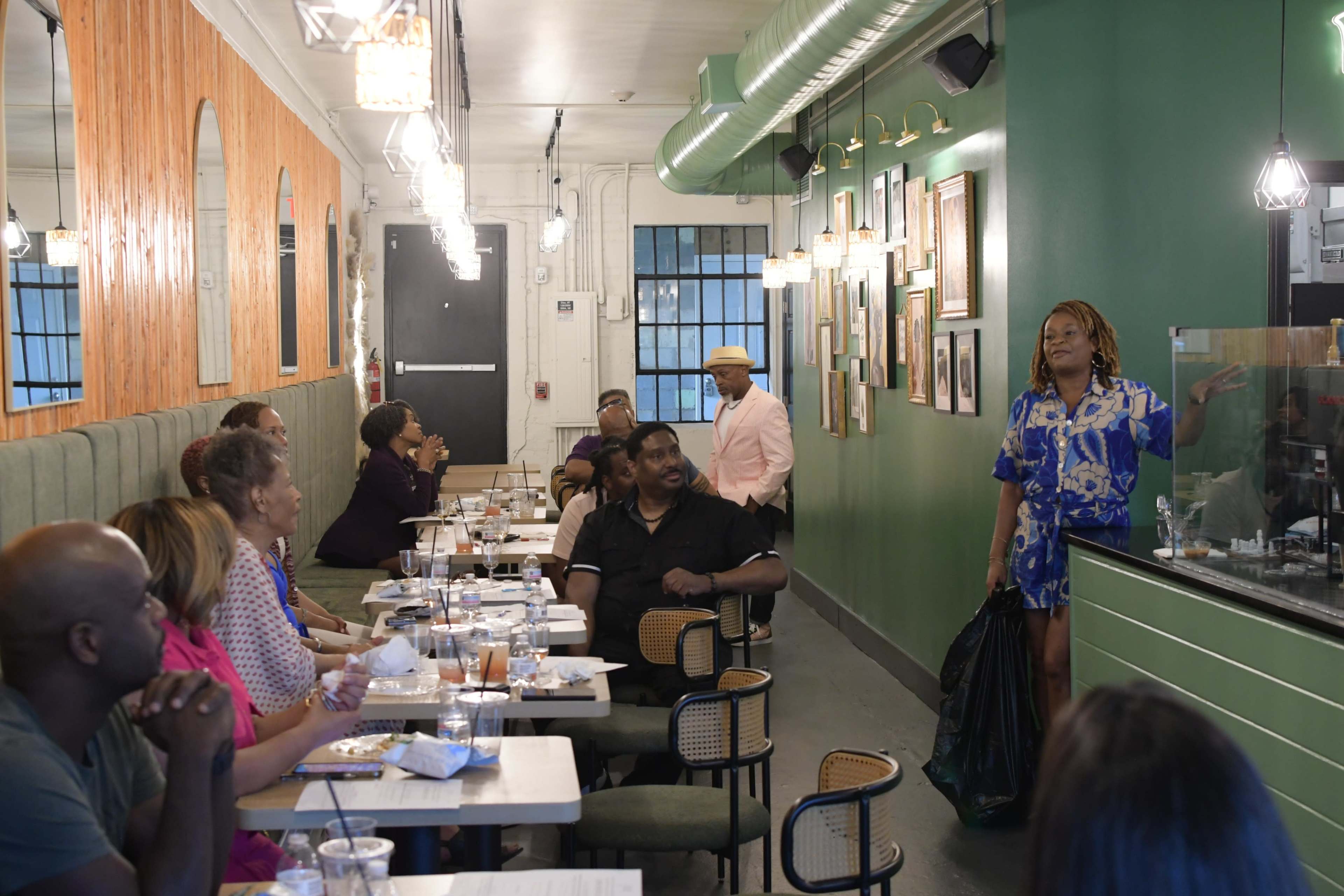 A woman wearing a blue patterned dress stands by a counter in a bustling restaurant, addressing a seated audience while others enjoy their meals at tables.