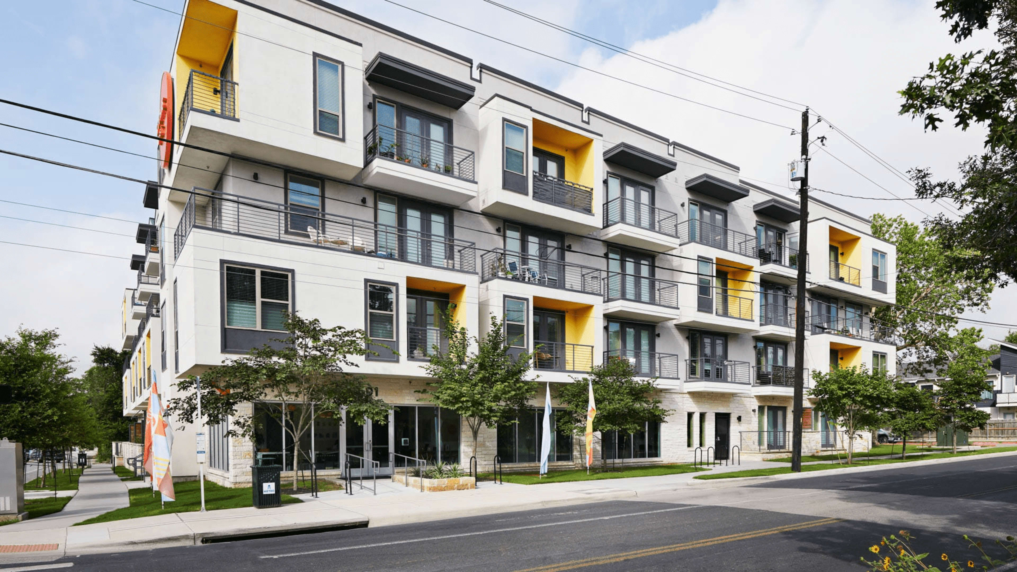The image shows a modern, multi-story residential building with balconies and large windows, surrounded by greenery and situated along a city street.