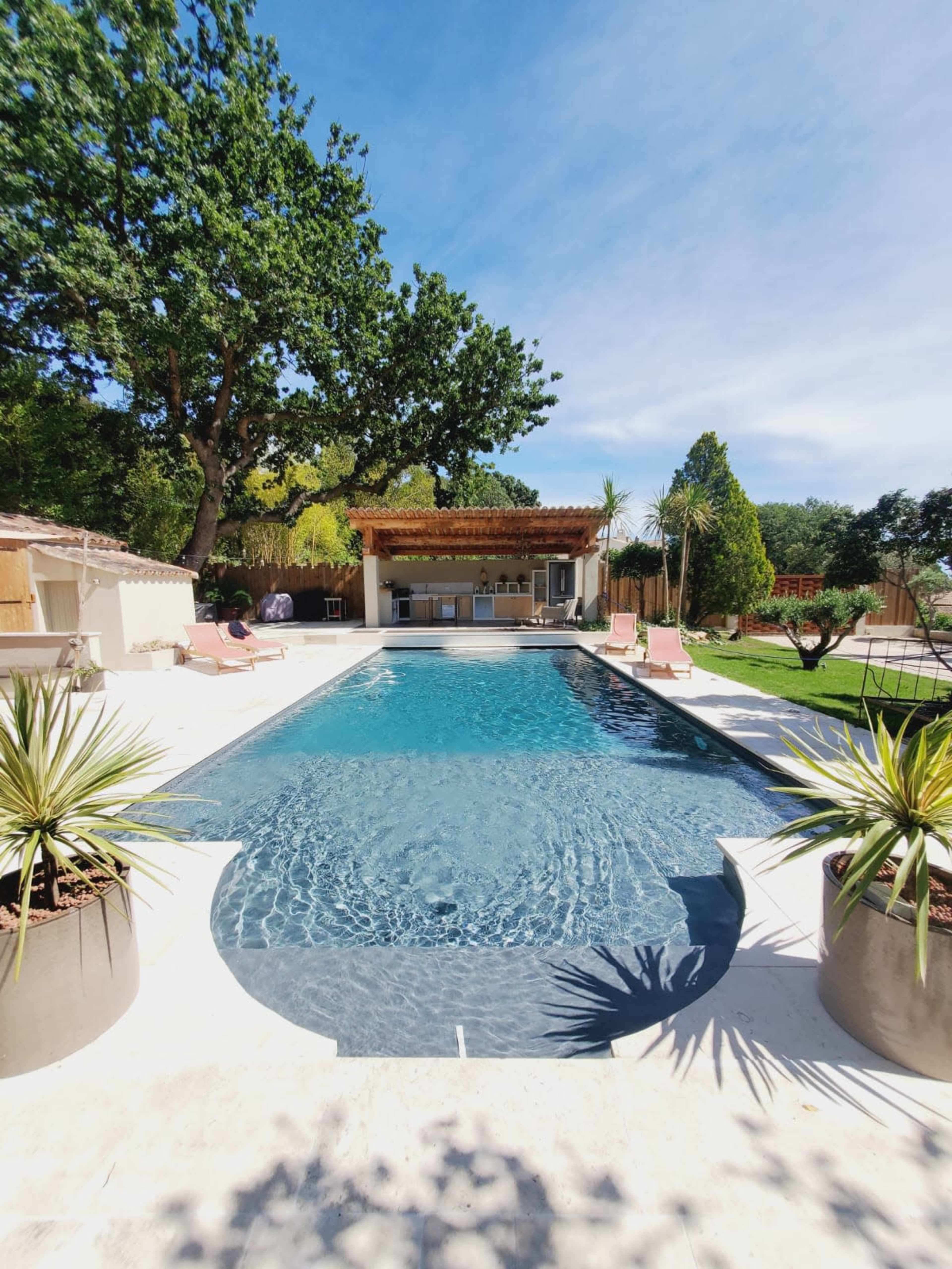 A rectangular swimming pool surrounded by potted plants and lounge chairs, with a covered outdoor kitchen and trees in the background.