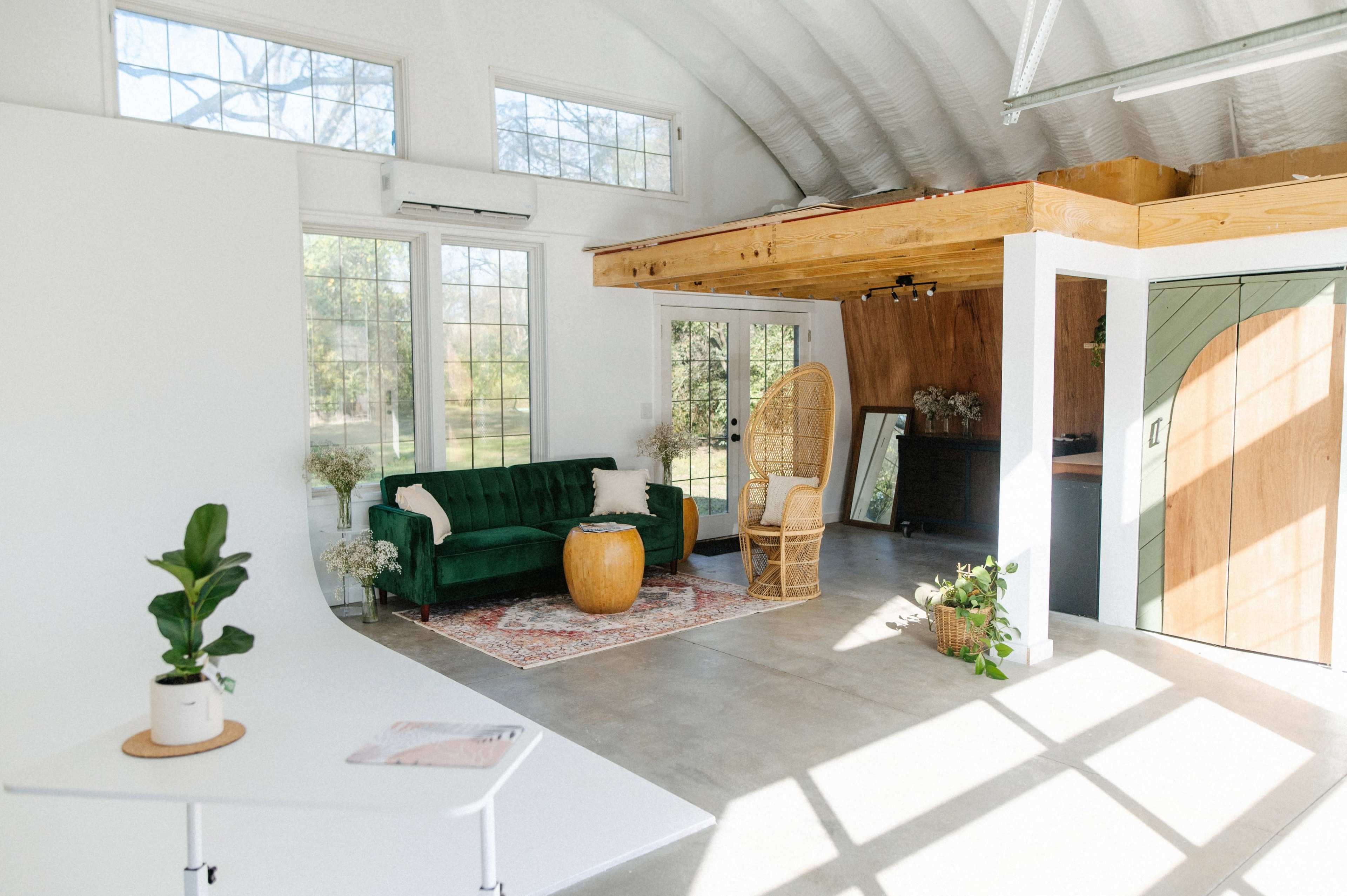 A bright, modern interior featuring a green sofa, a rattan chair, and large windows allowing natural light to fill the space.