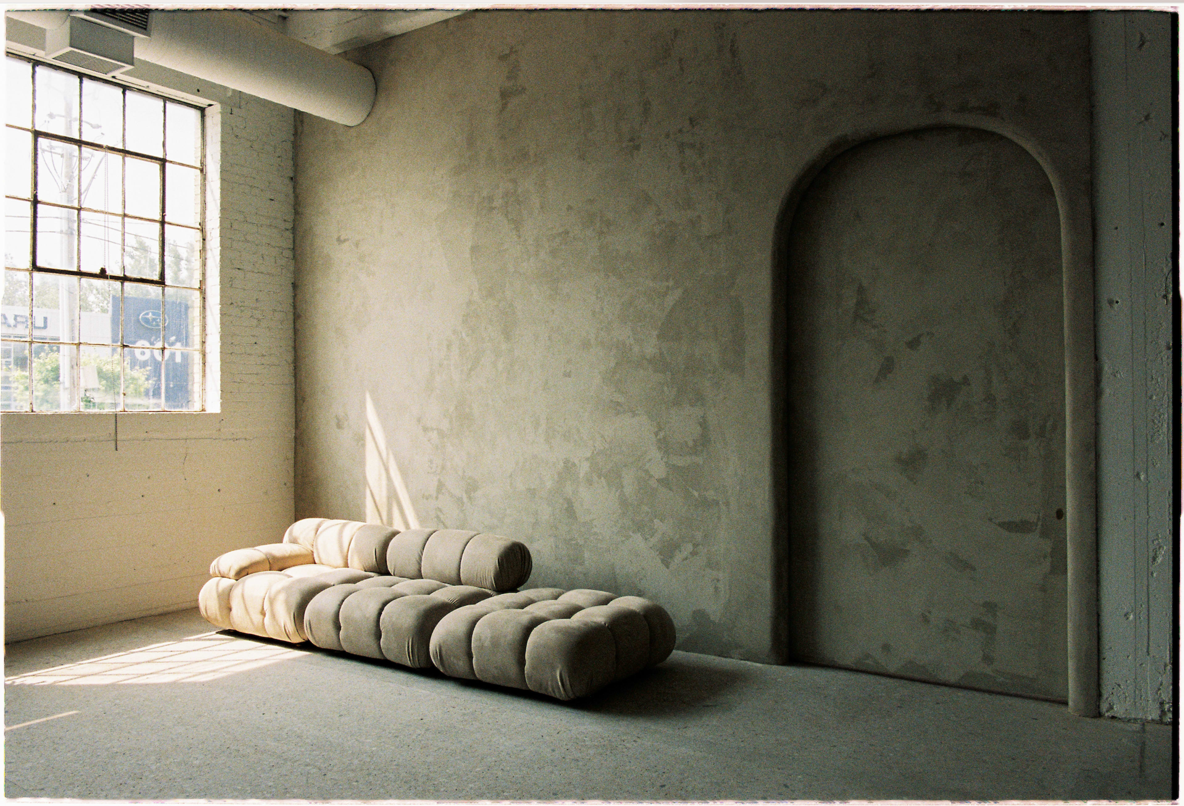 A minimalist interior space with a beige cushioned sofa positioned against a textured gray wall and a large window letting in natural light.