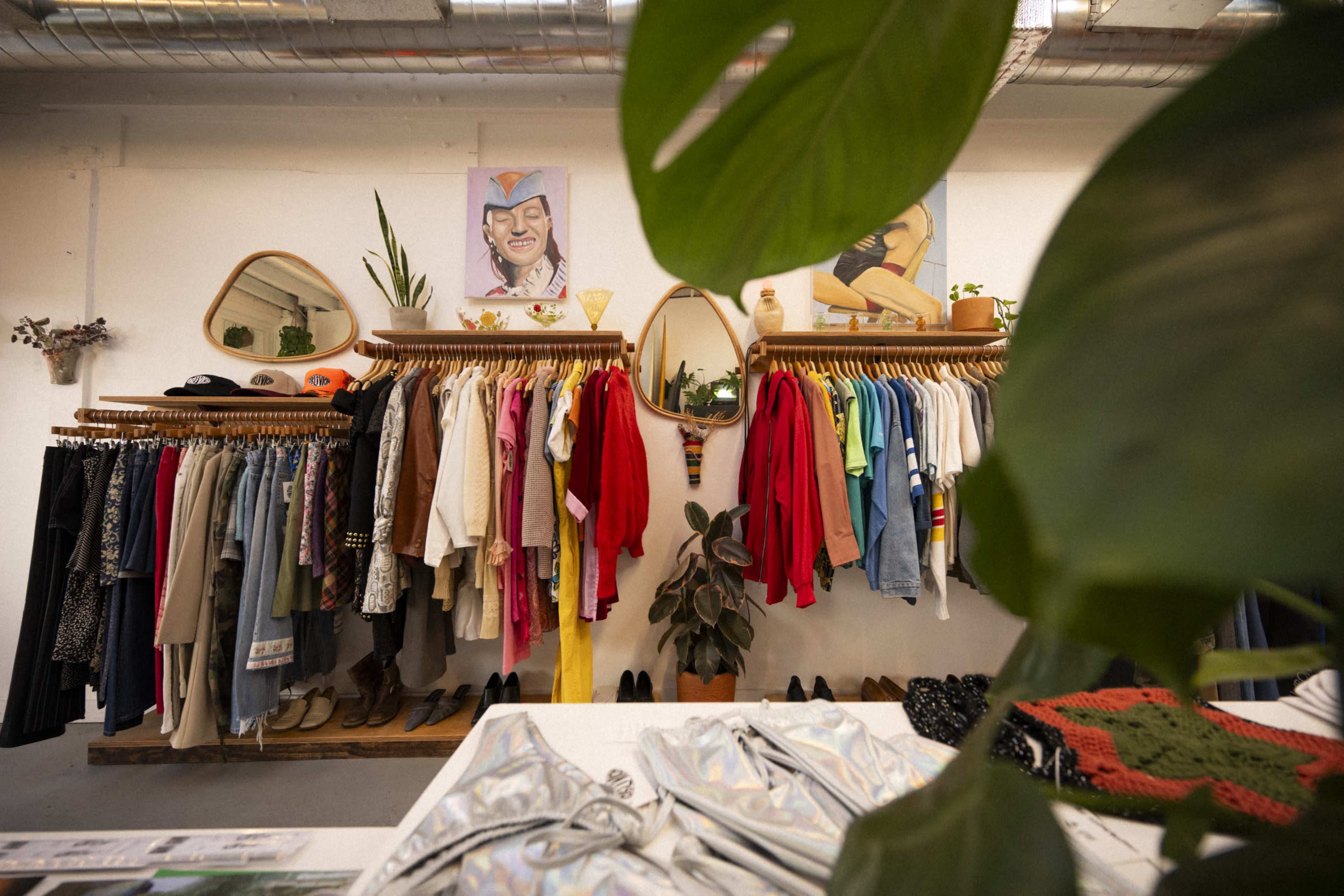 A clothing store interior features racks of various garments, including shirts and jackets, with decorative plants and artwork on the walls.