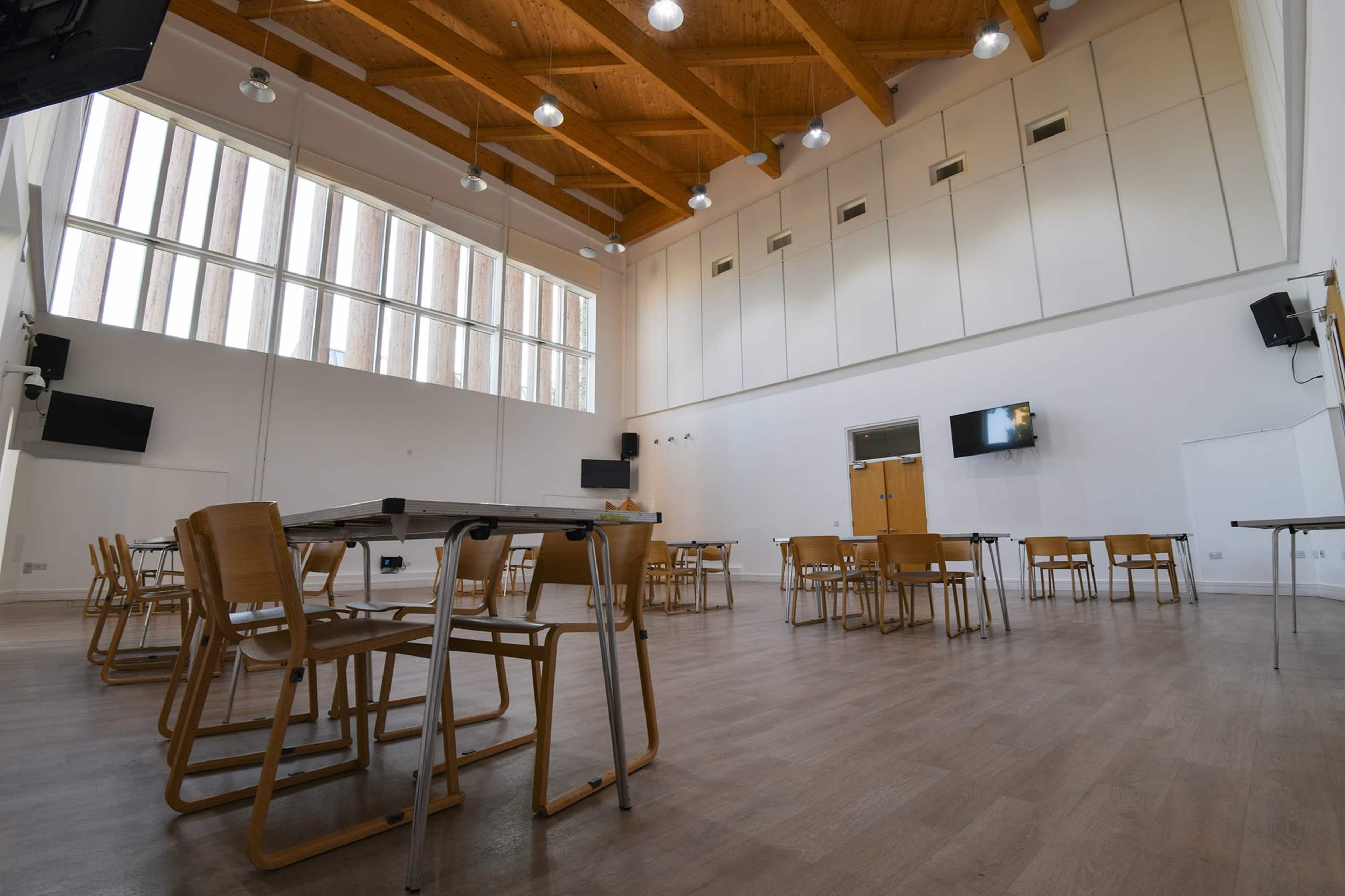 A spacious meeting room features wooden beams, several tables, and chairs arranged on a light-colored floor.