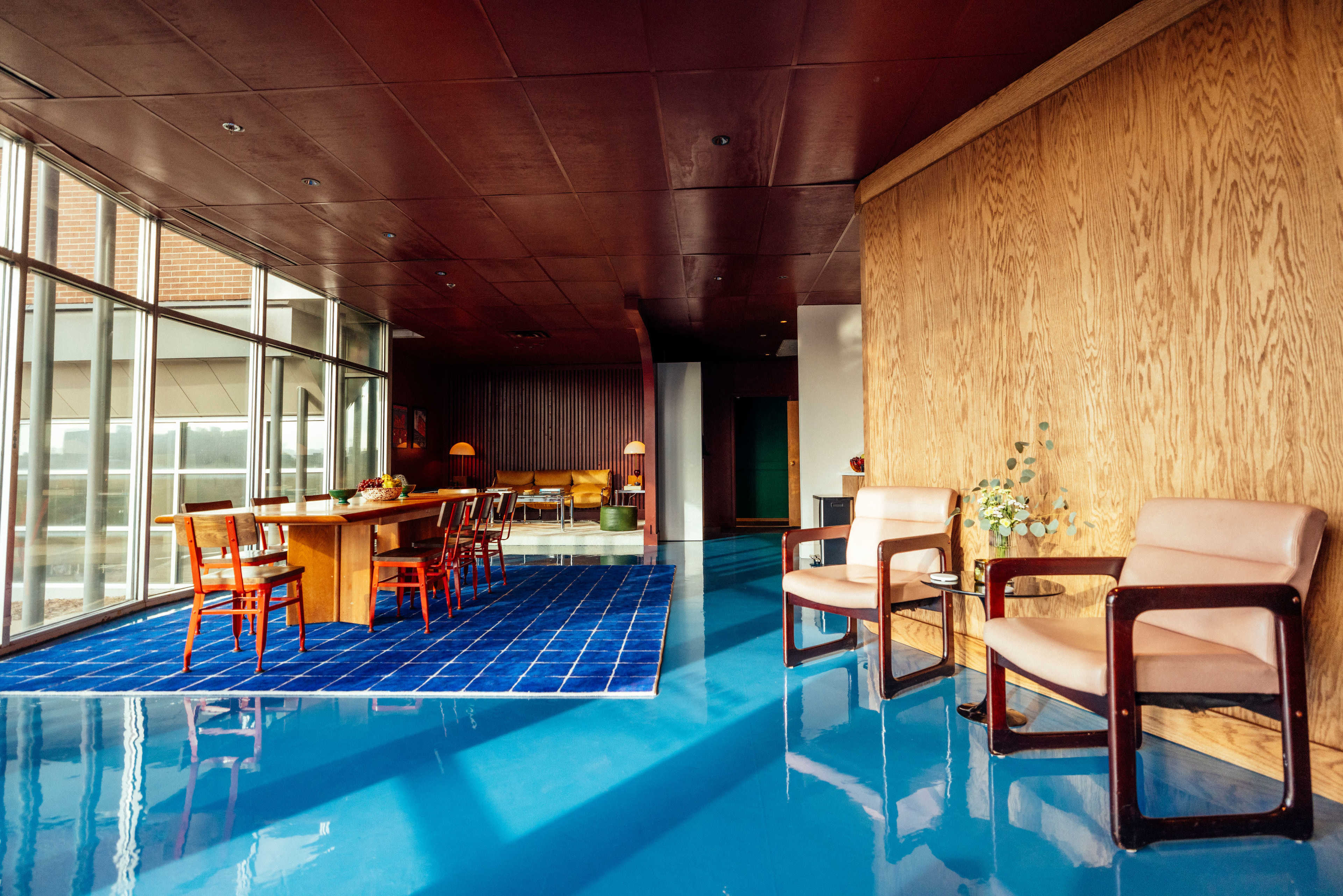 The image shows a modern interior space featuring a dining area with wooden furniture, a blue rug, large windows, and a polished blue floor.
