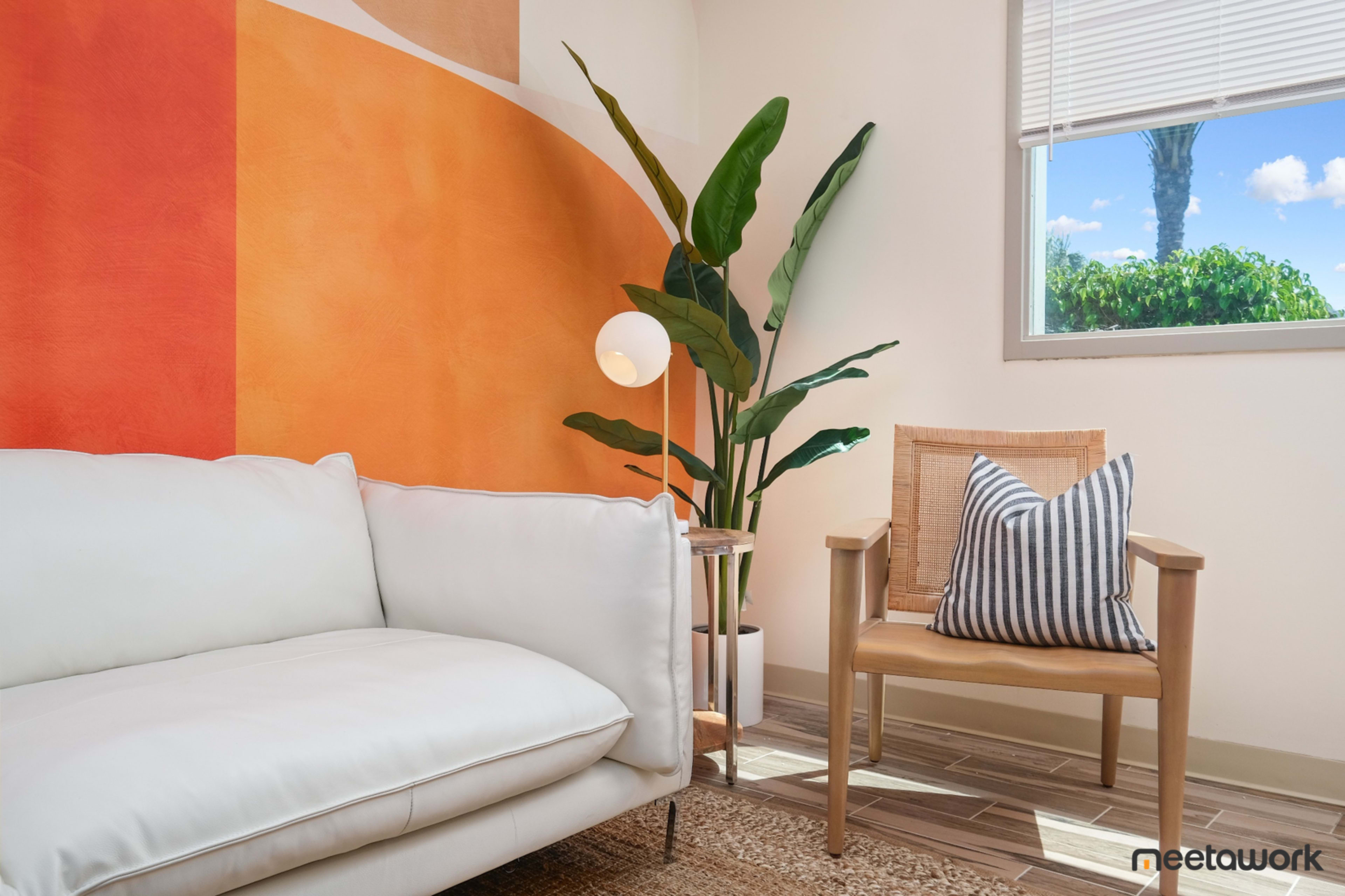 A bright interior scene featuring a white couch, a rattan chair with a striped pillow, a floor lamp, and a large indoor plant next to an orange accent wall.