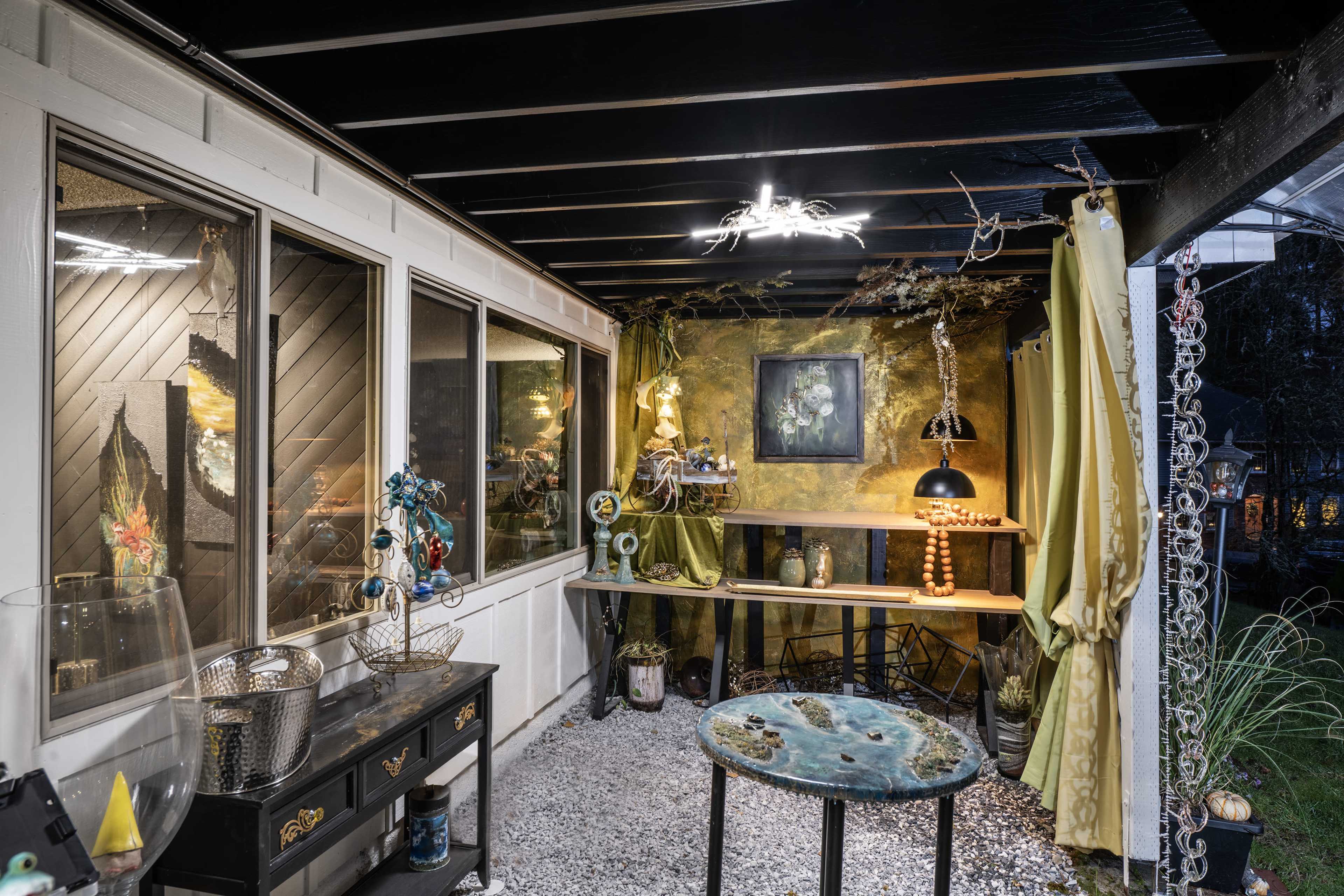 The image shows a covered outdoor space featuring various decorative items, a table, and art displayed on the walls.