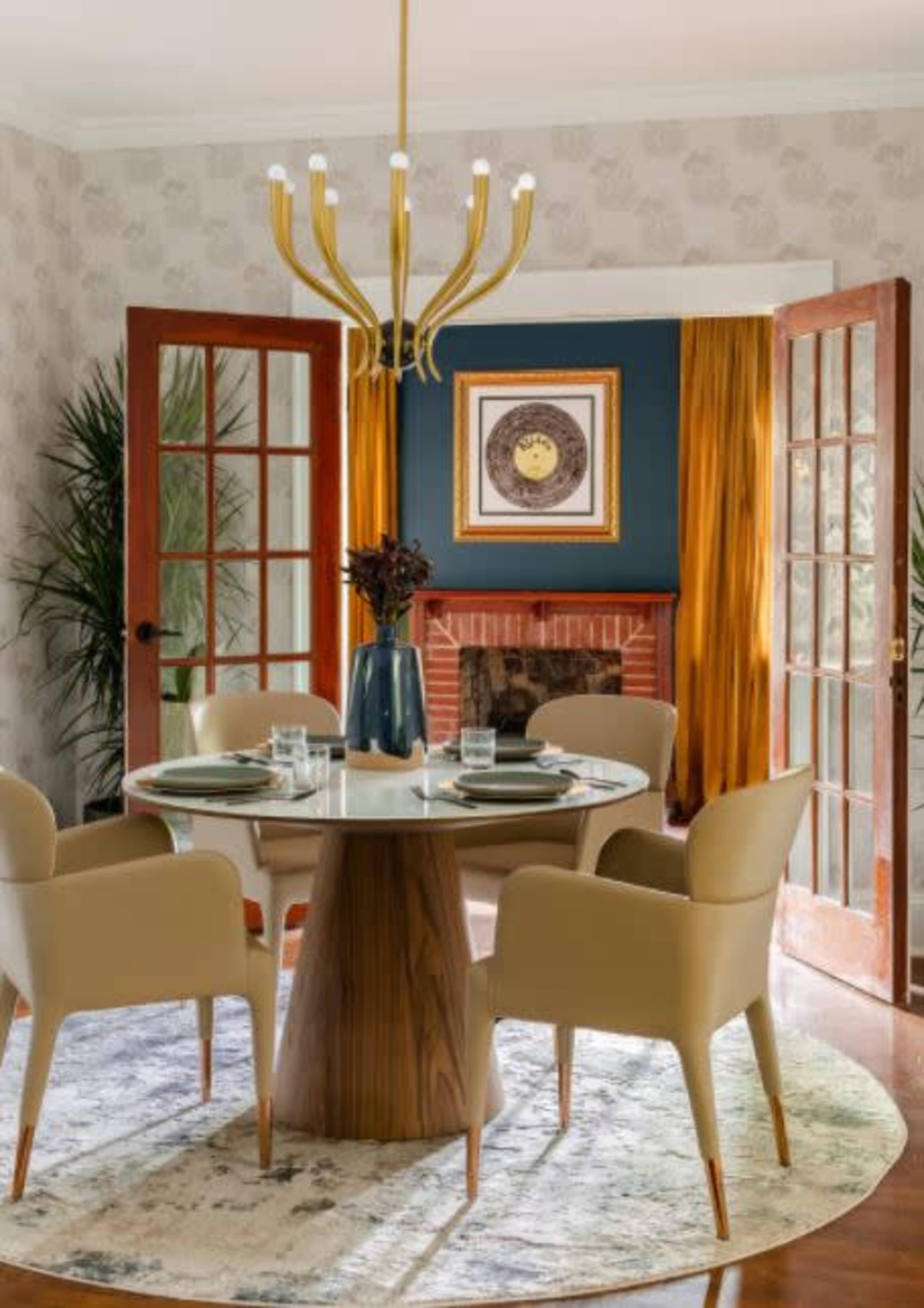 A modern dining room features a round table surrounded by four beige chairs, with a chandelier overhead and a fireplace visible through double doors.