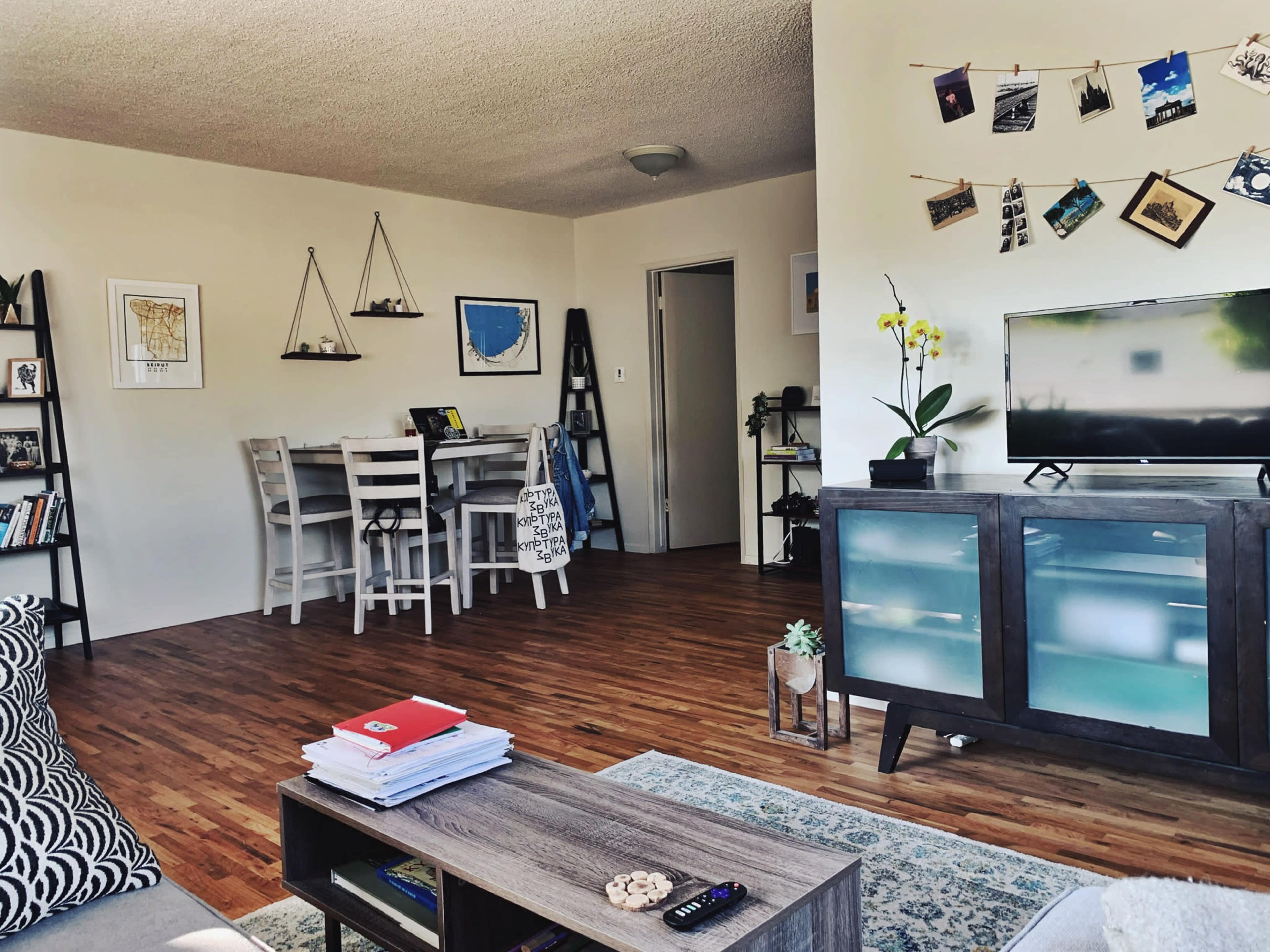 The image shows a living room with wooden flooring, a television on a cabinet, a dining area with white chairs, and various decorations on the walls.