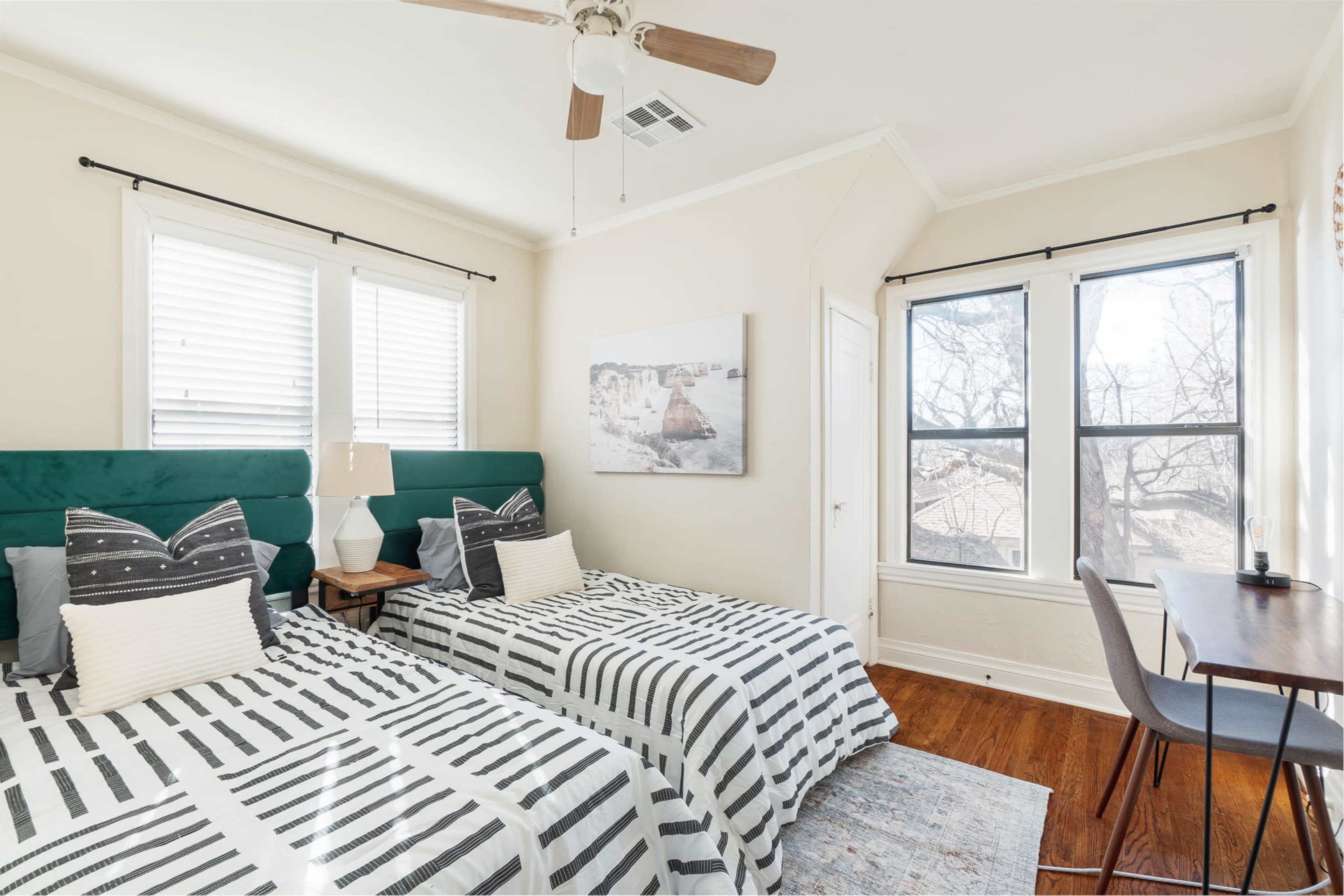A bedroom featuring two twin beds with striped bedding, a wooden nightstand, a ceiling fan, and large windows with natural light.