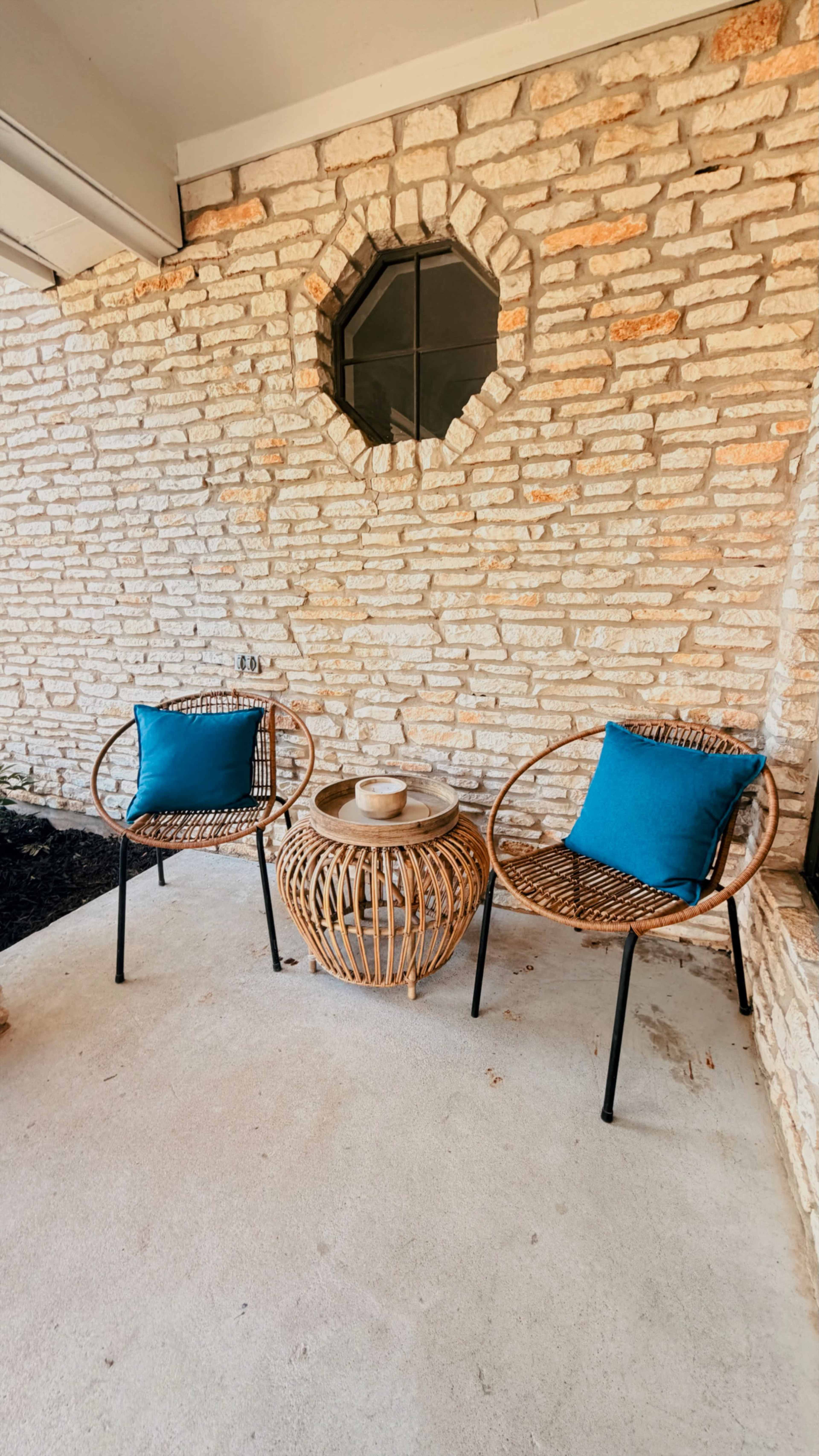Two rattan chairs with blue cushions are positioned around a round, woven table on a concrete porch next to a brick wall with an octagonal window.