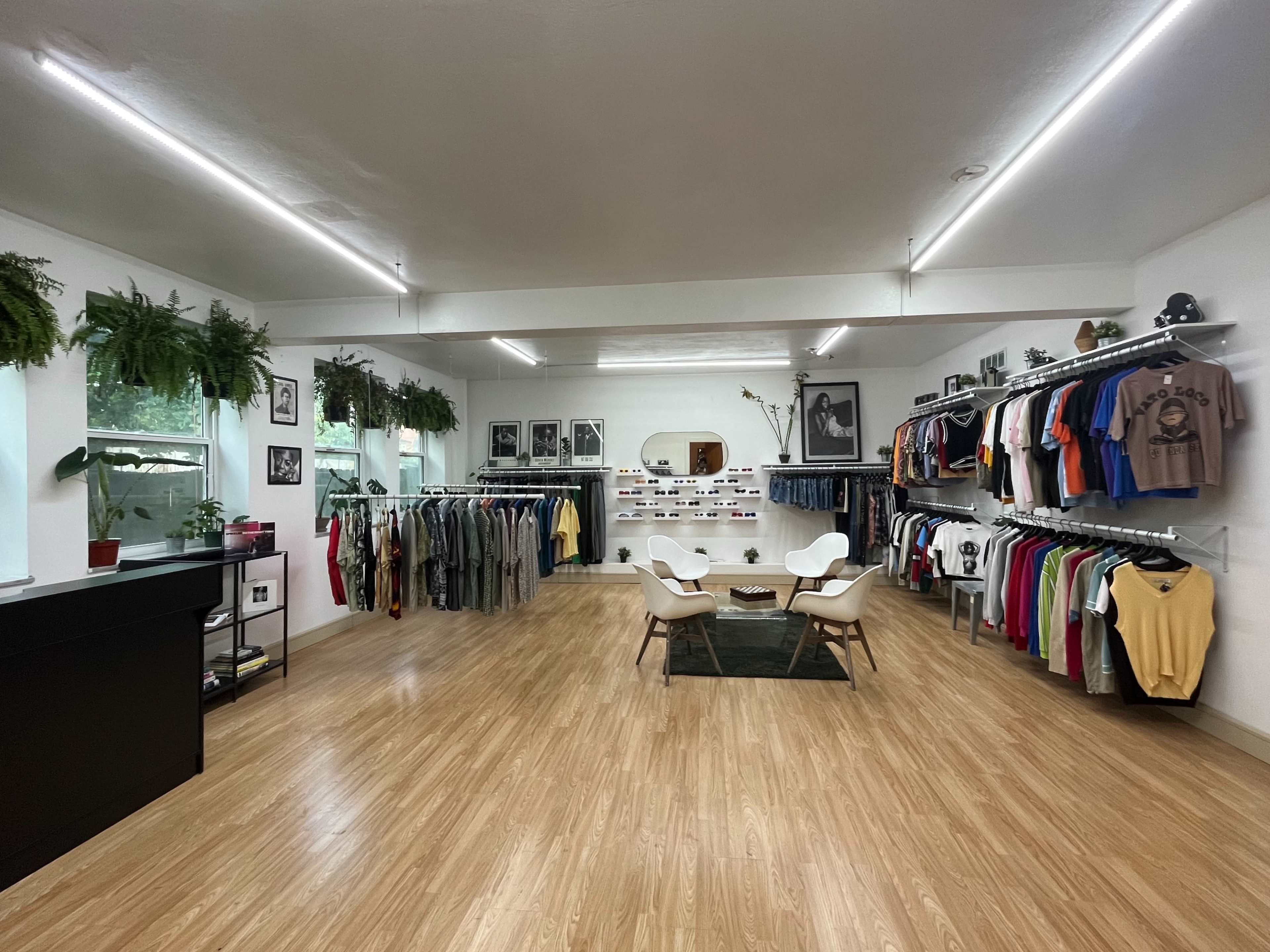 A modern clothing store features rows of hanging apparel, minimalist furniture, and decor elements like plants and framed photographs.