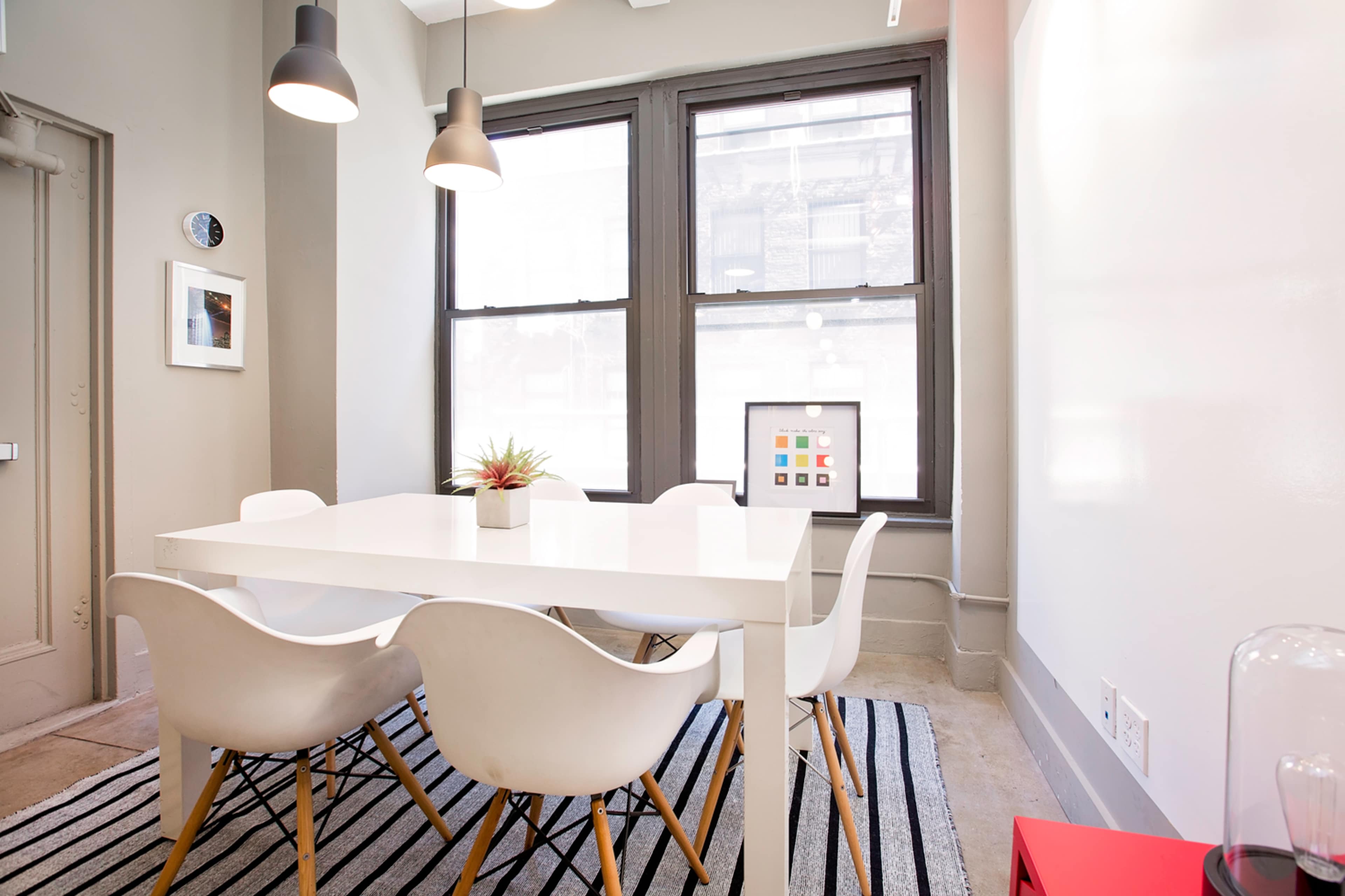 A modern dining area features a white table surrounded by six white chairs, with large windows and a patterned rug on the floor.