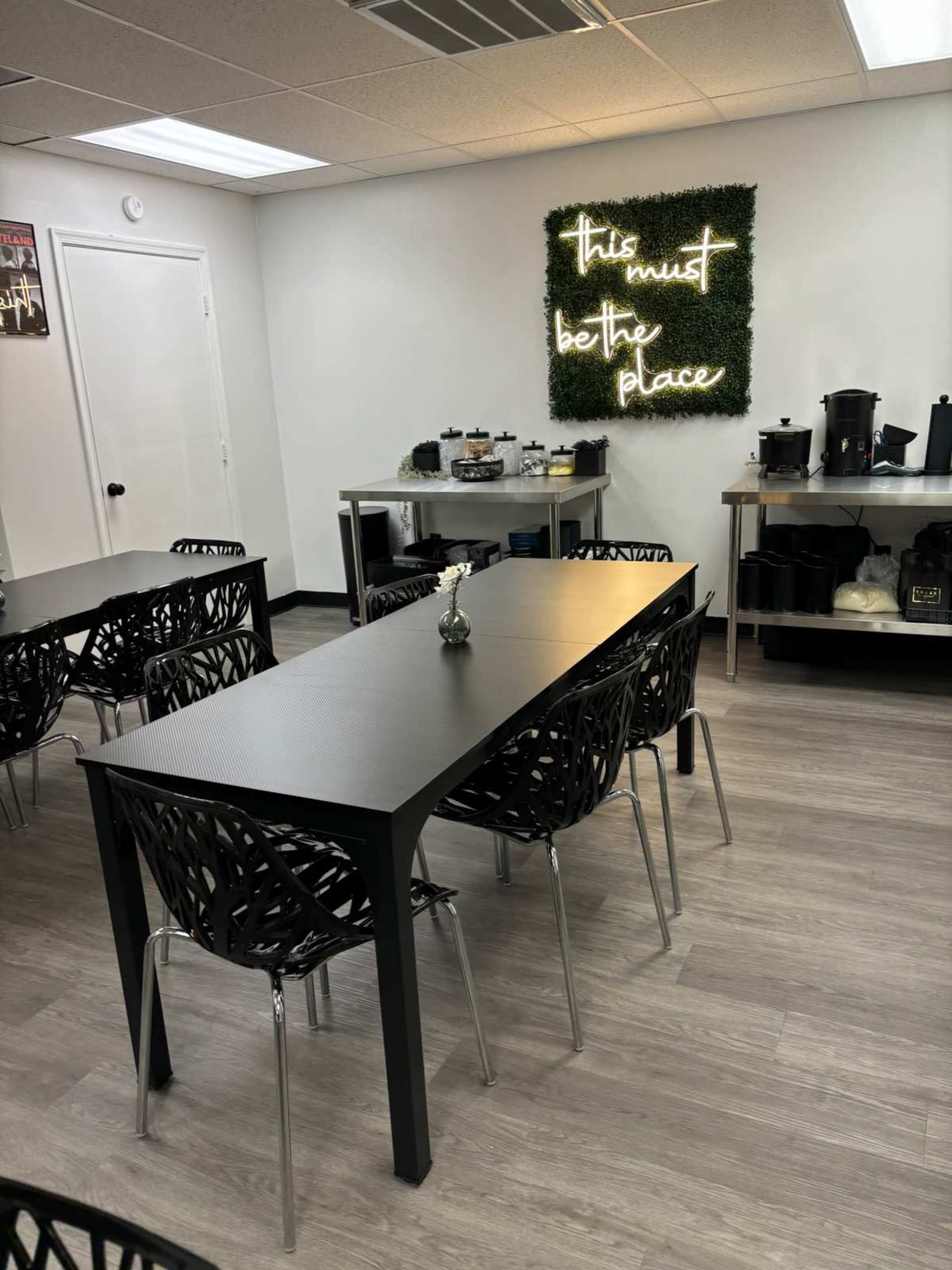 The image shows a modern break room with black tables and chairs, featuring a neon sign that reads "this must be the place" on a green wall.