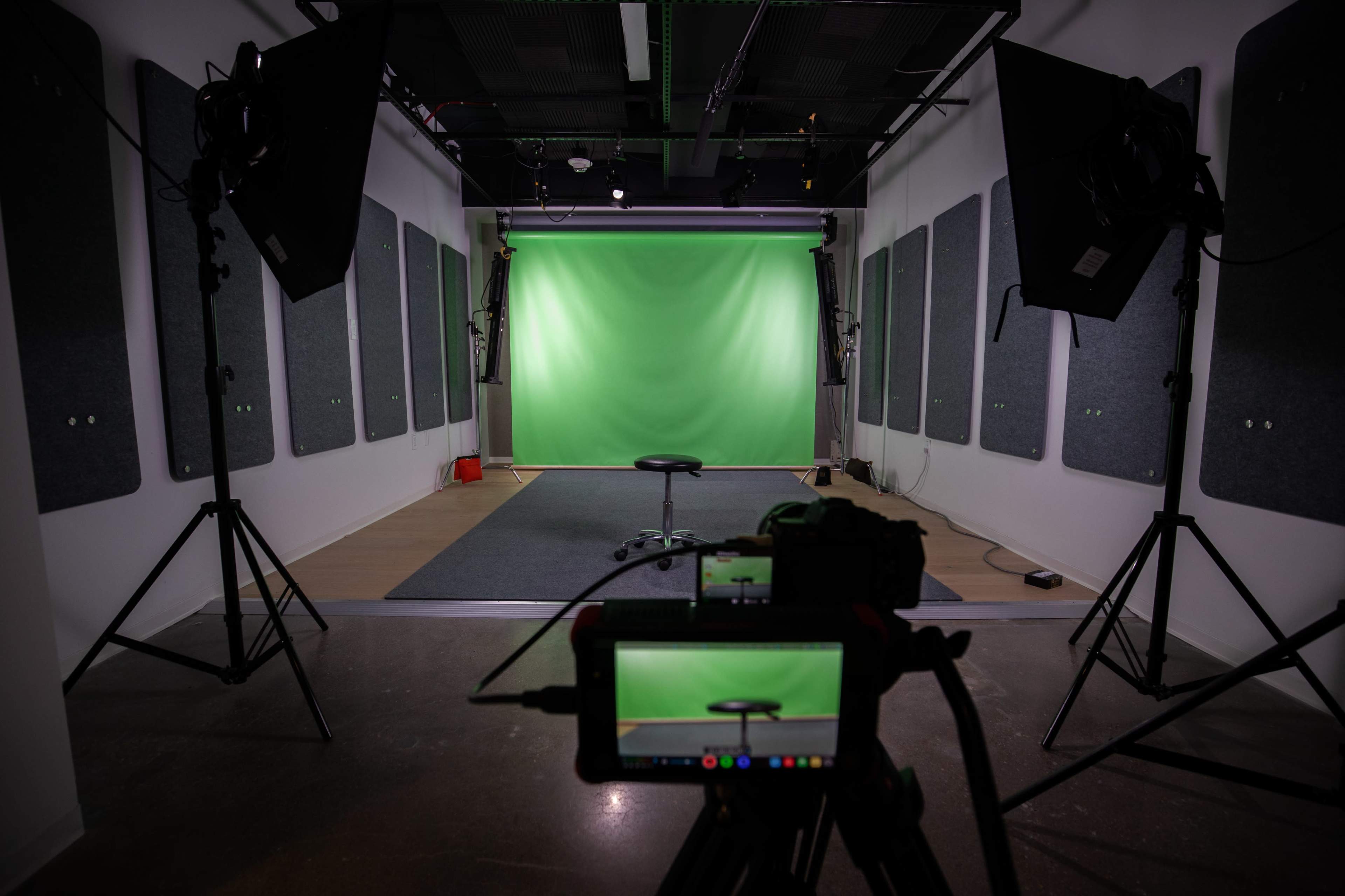 A studio setup with a green screen backdrop, a black stool in the center, and professional lighting equipment on either side.