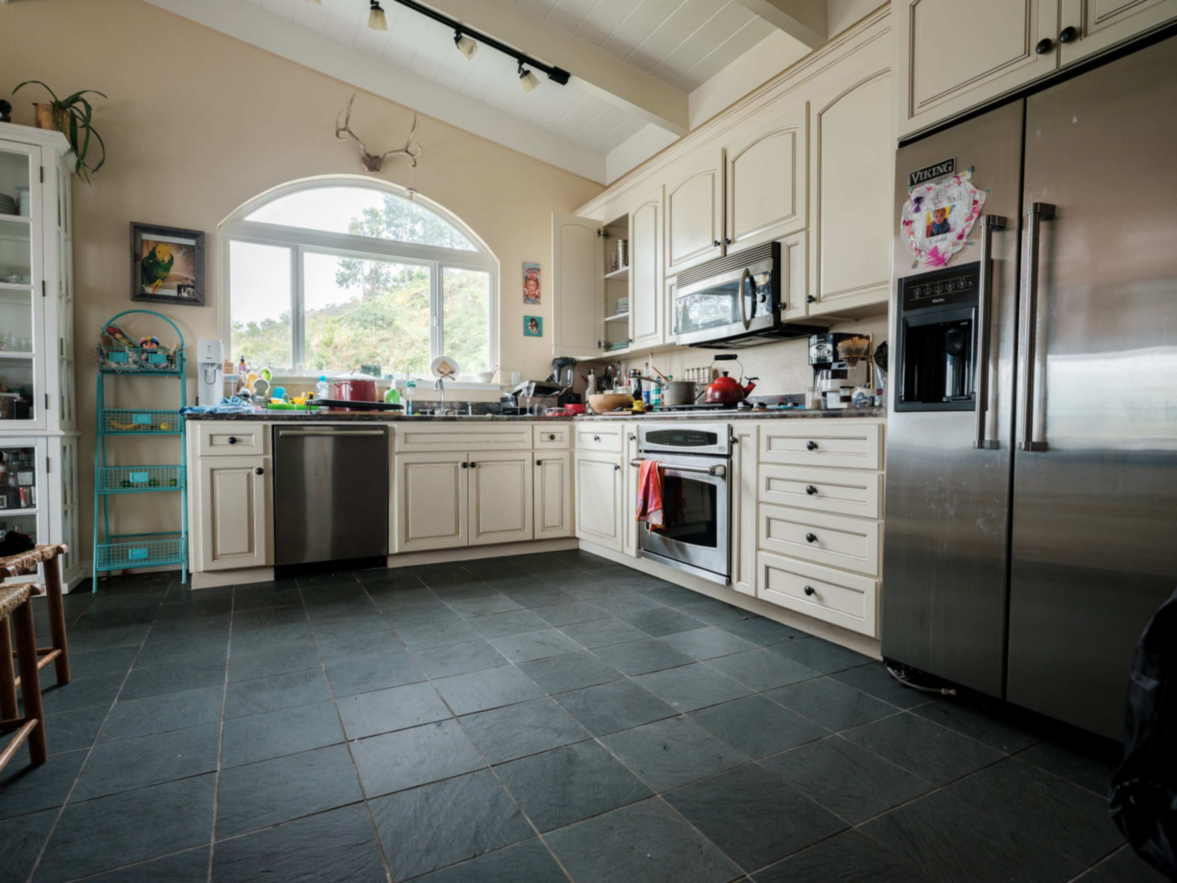 A spacious kitchen features cream-colored cabinets, stainless steel appliances, and a large window overlooking a scenic view, with various cooking items scattered across the countertops.
