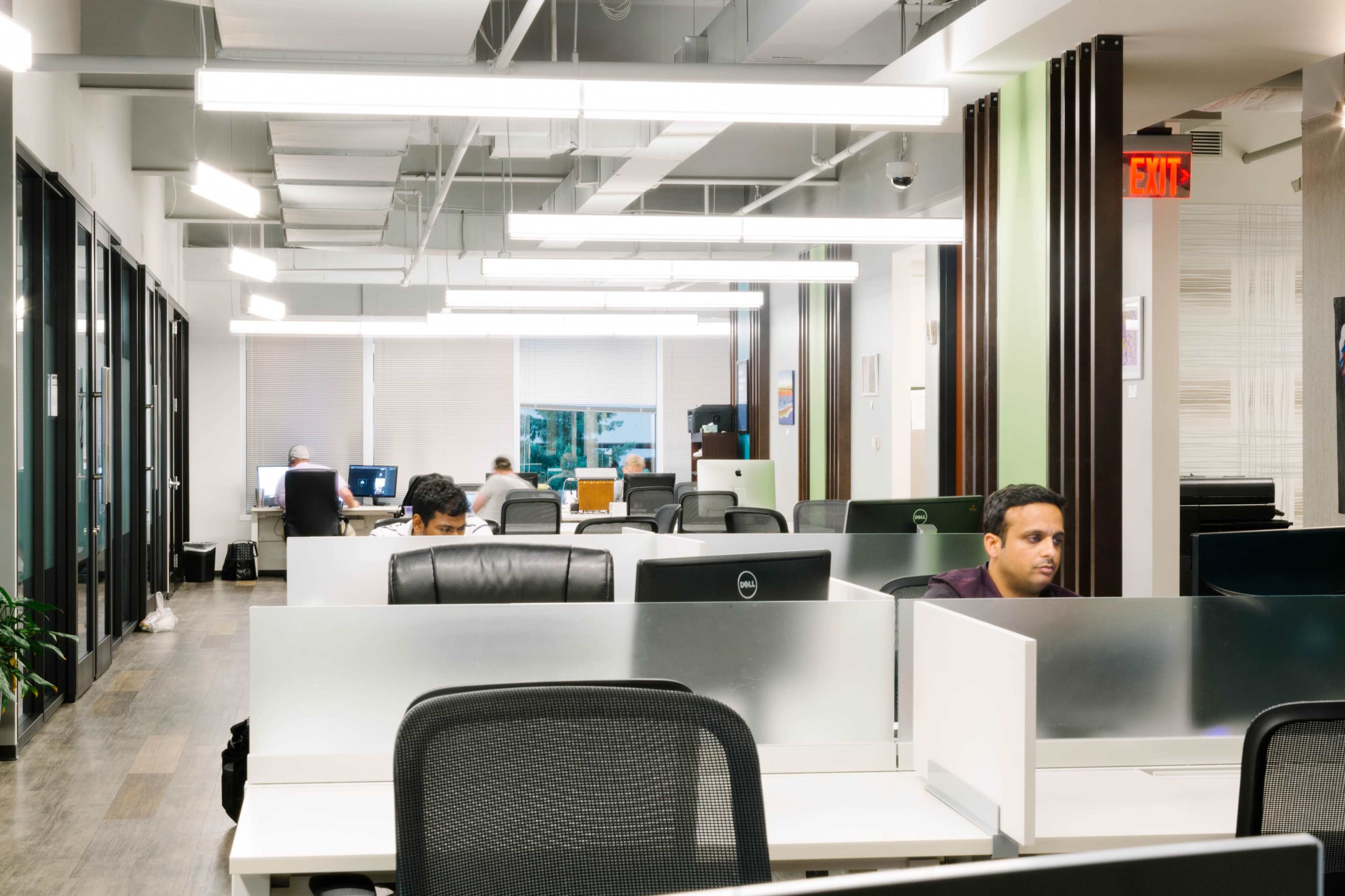 An open office space features several workstations with people engaged in computer tasks, illuminated by overhead lights.