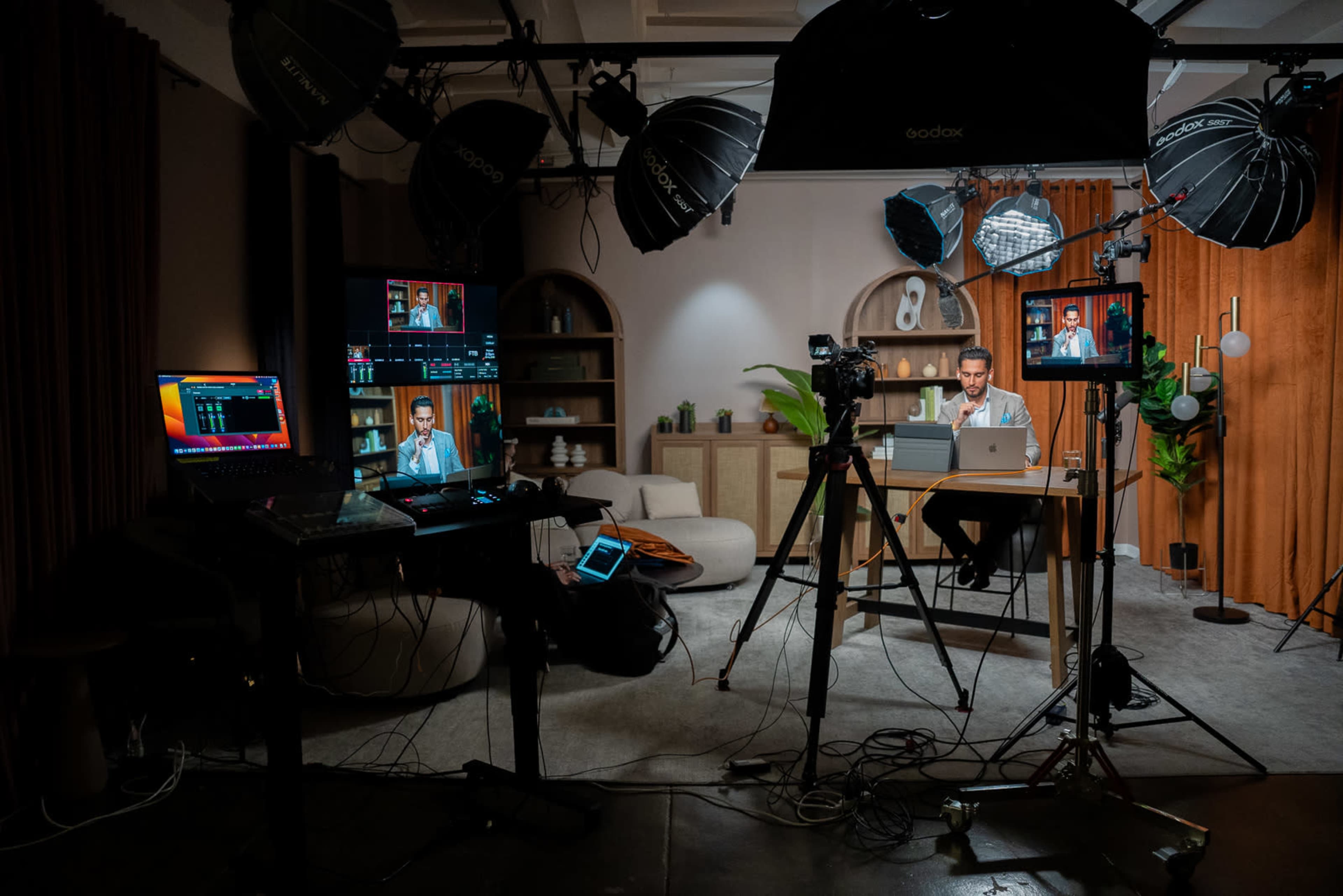 The image shows a well-equipped studio set up for video production, featuring multiple cameras, lights, and screens, with a presenter sitting at a desk.