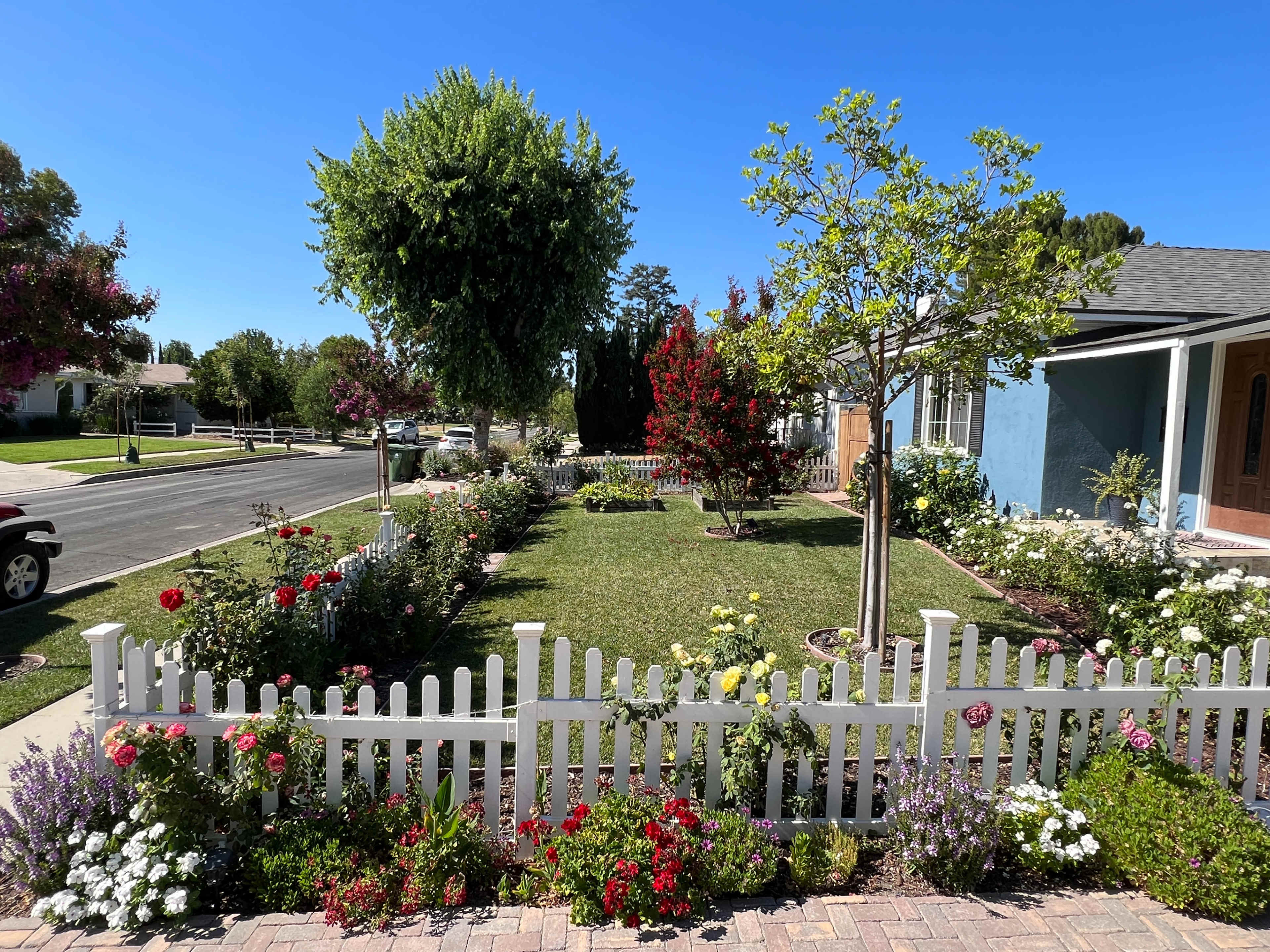 A white picket fence encloses a landscaped yard with flowerbeds, trees, and a manicured lawn alongside a quiet street.
