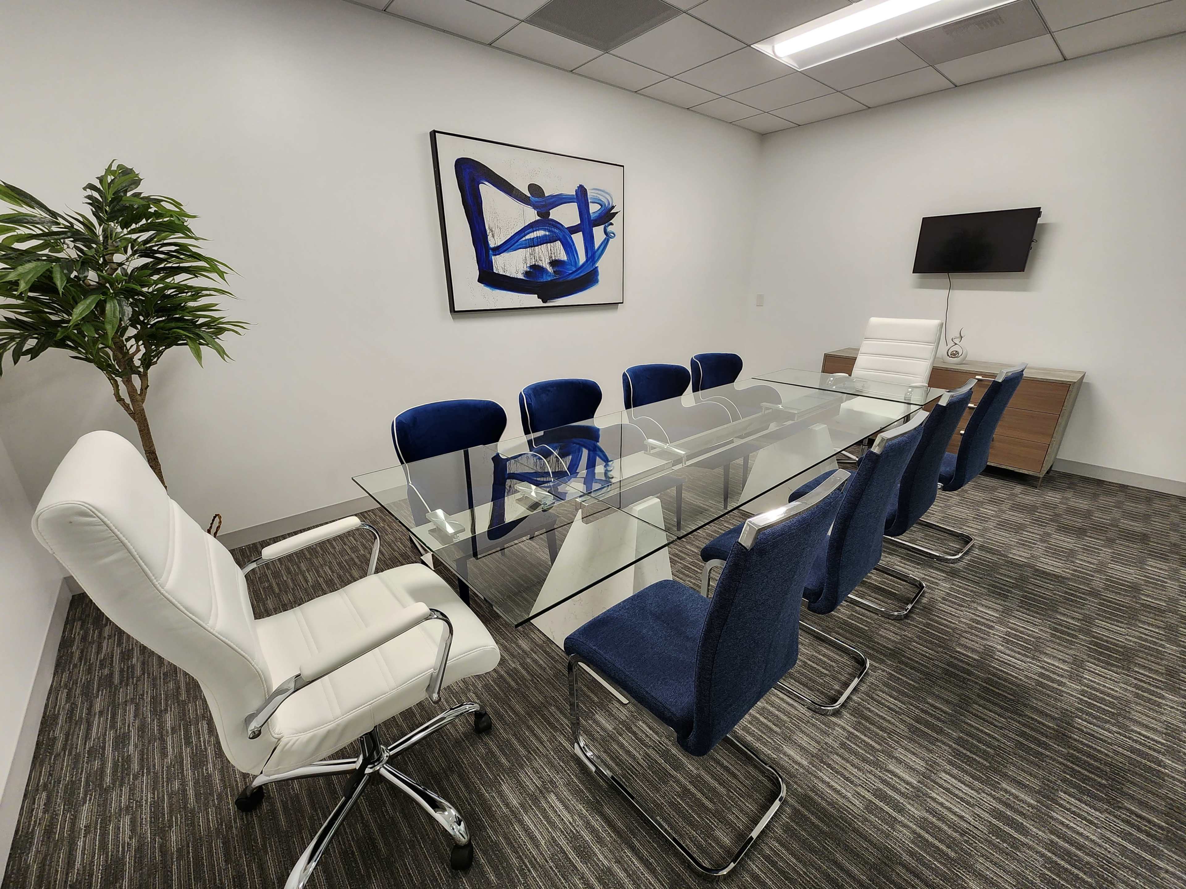 A modern conference room features a glass table surrounded by blue upholstered chairs and a white leather chair, with a large abstract painting on the wall.