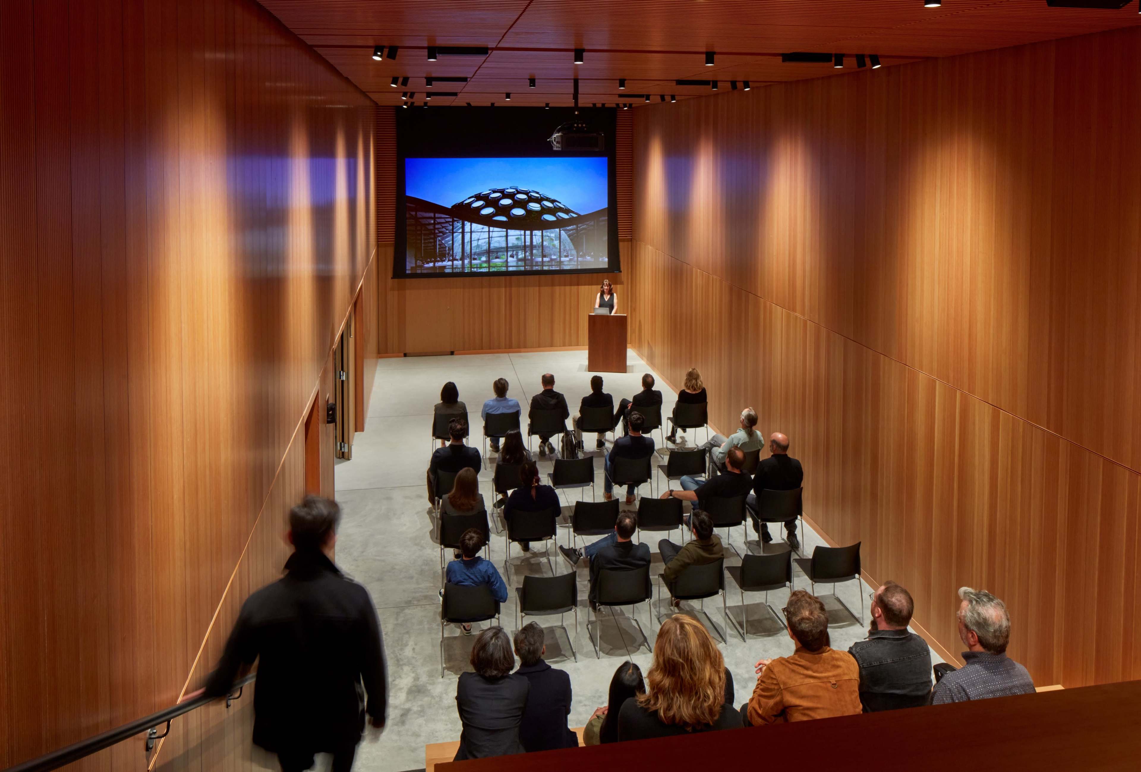 A speaker presents to an audience in a modern auditorium with wooden walls and a large screen displaying images.