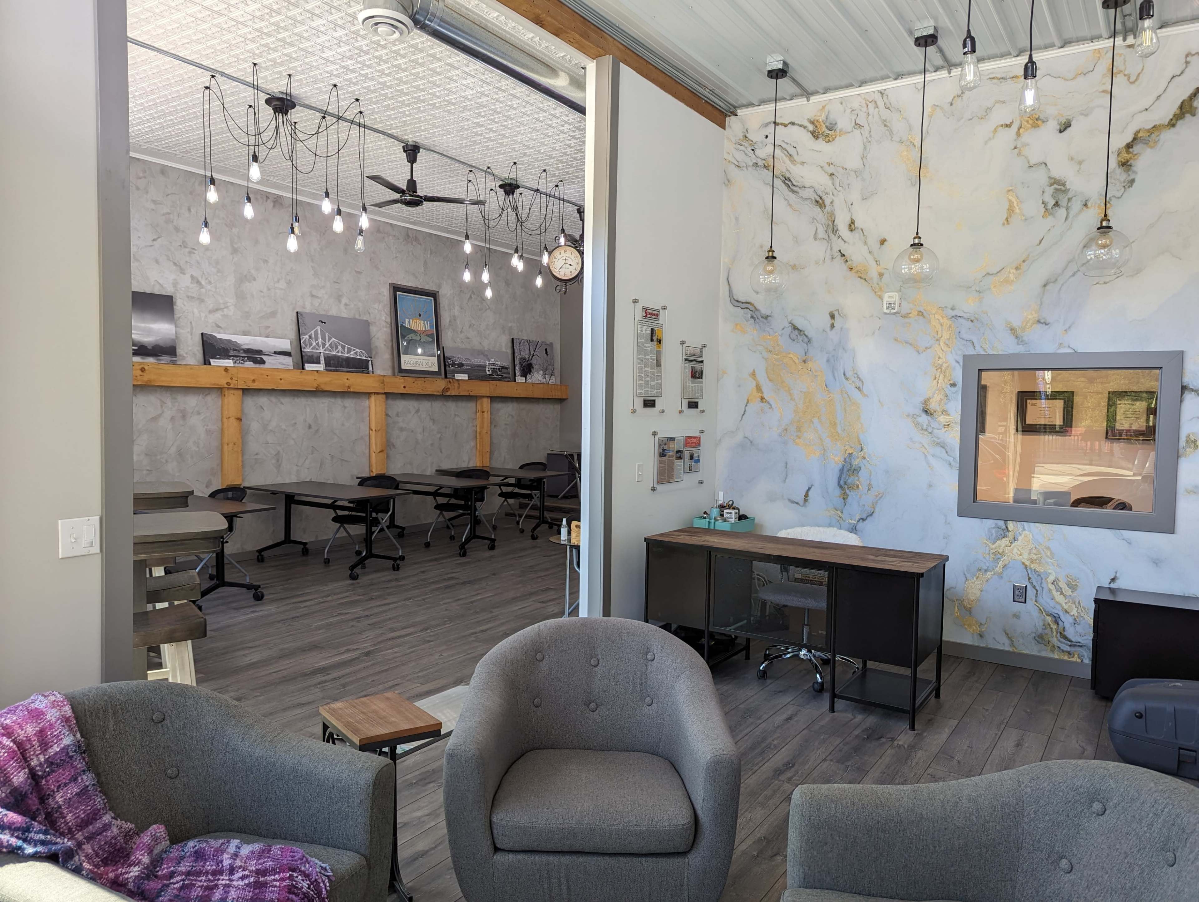 A modern interior space with a seating area featuring gray armchairs and a desk, leading to a classroom-like area with tables and a decorative marble wall.