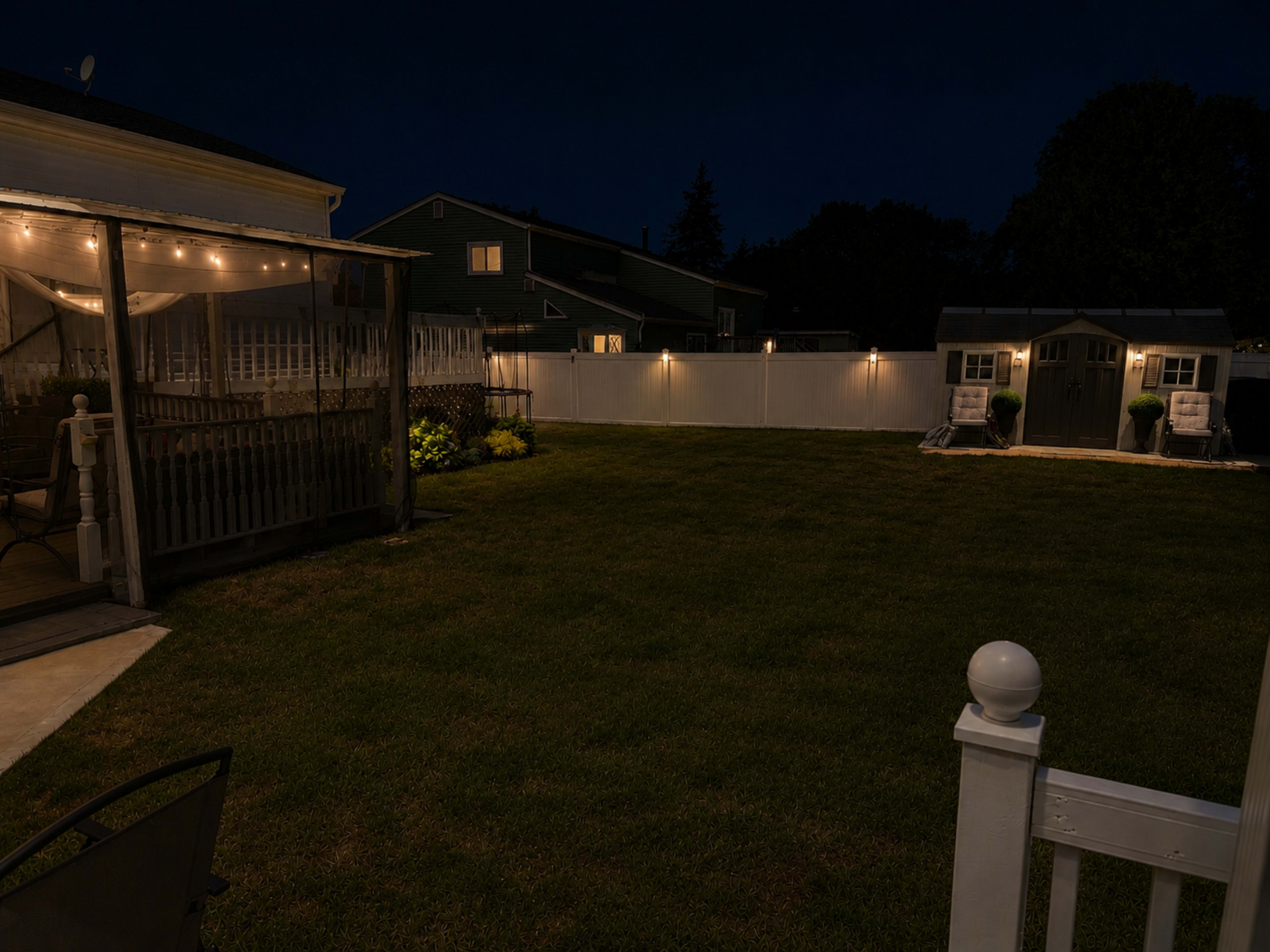 The image shows a well-maintained backyard at night, featuring a grassy area, a lit patio, and a small shed against a white fence.