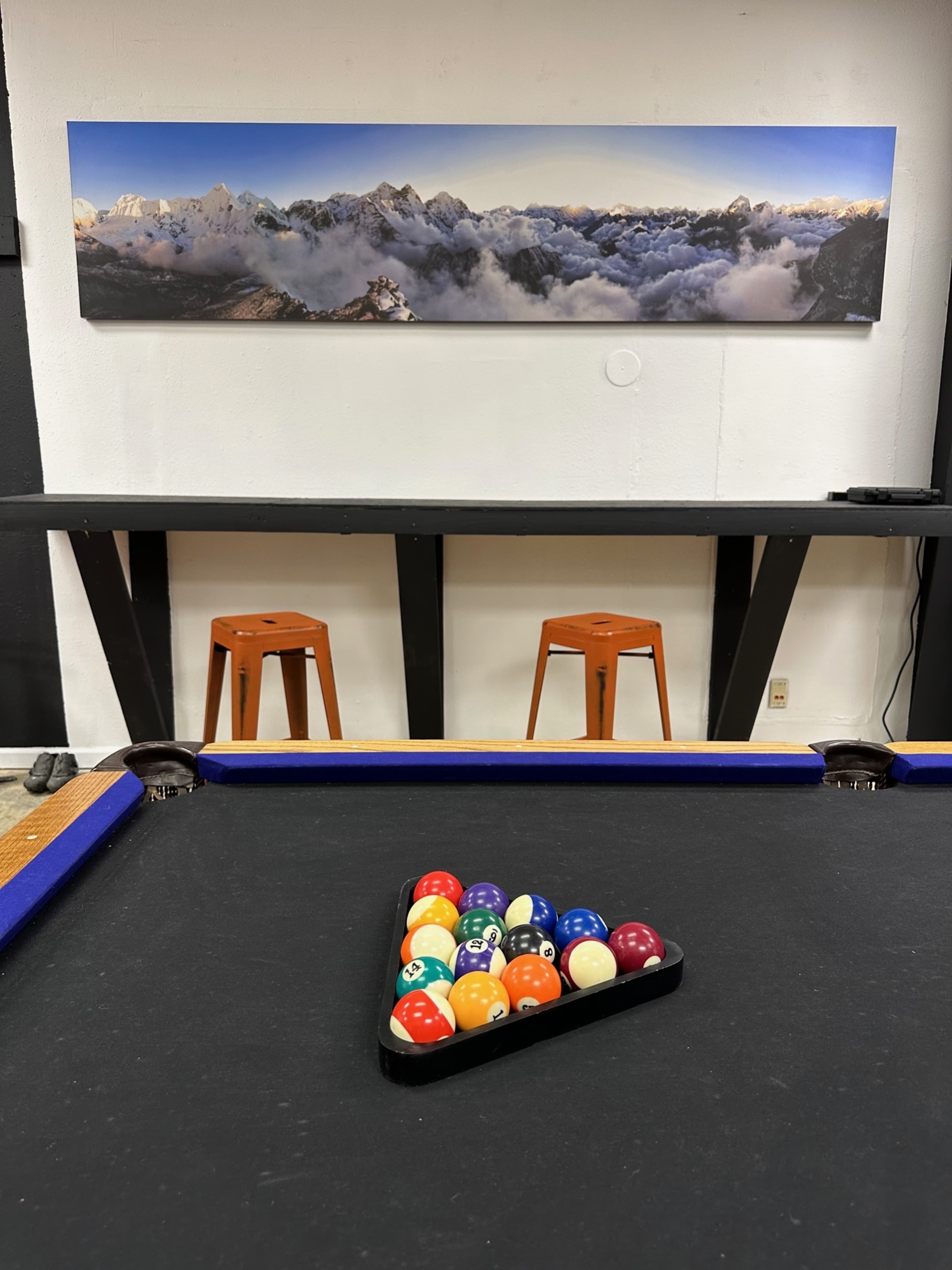 A pool table with a triangle rack of colorful billiard balls sits in front of a wall featuring a panoramic mountain landscape, with two orange stools positioned at a bar behind.