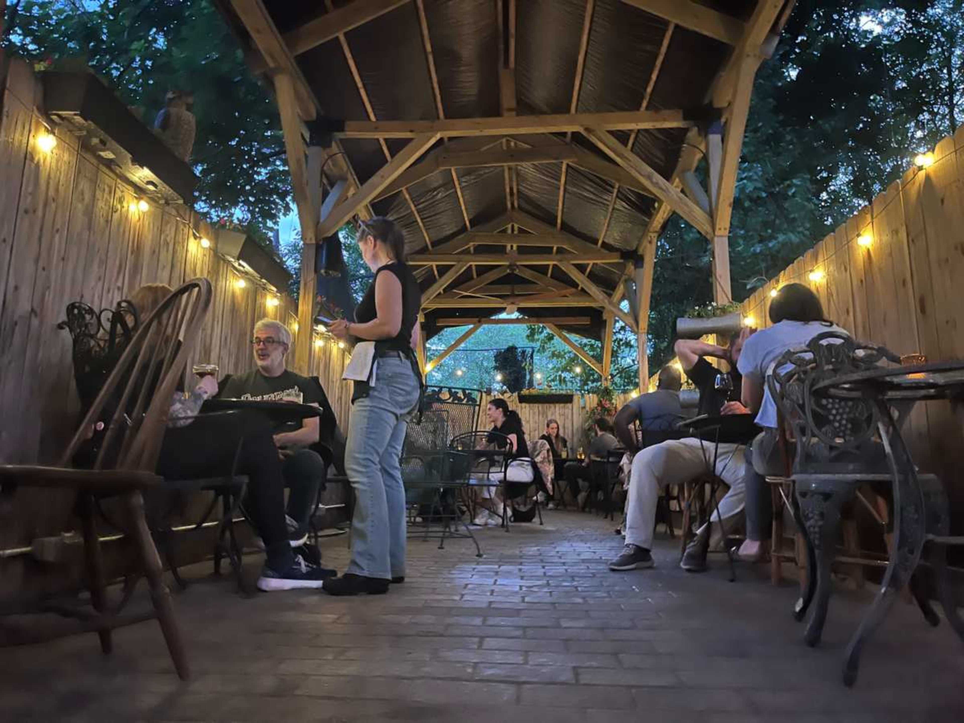 A dimly lit outdoor patio features several tables with patrons dining and chatting under string lights and a wooden roof.