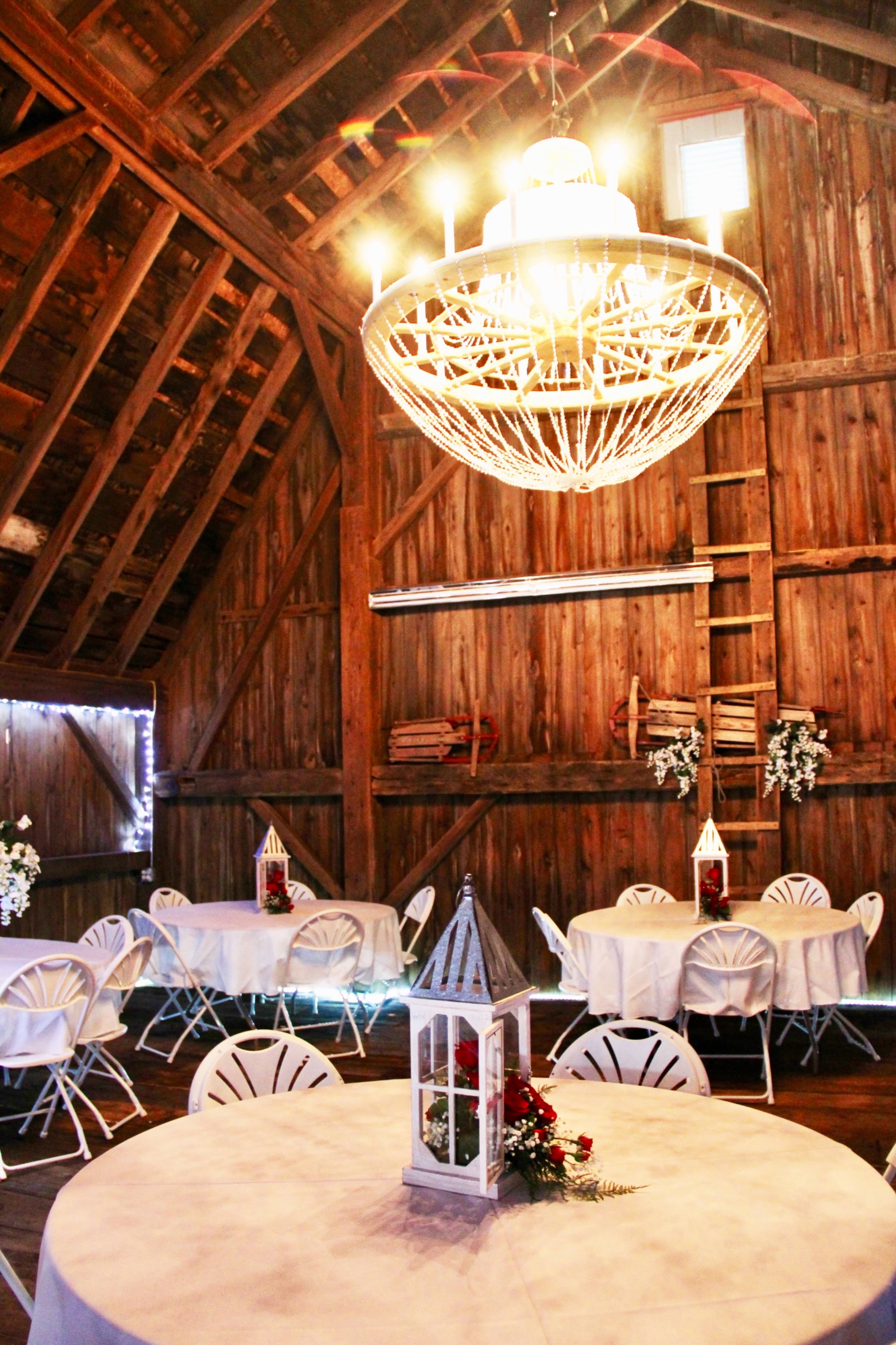 The interior of a wooden barn features round tables with white tablecloths and a large chandelier hanging from the ceiling.