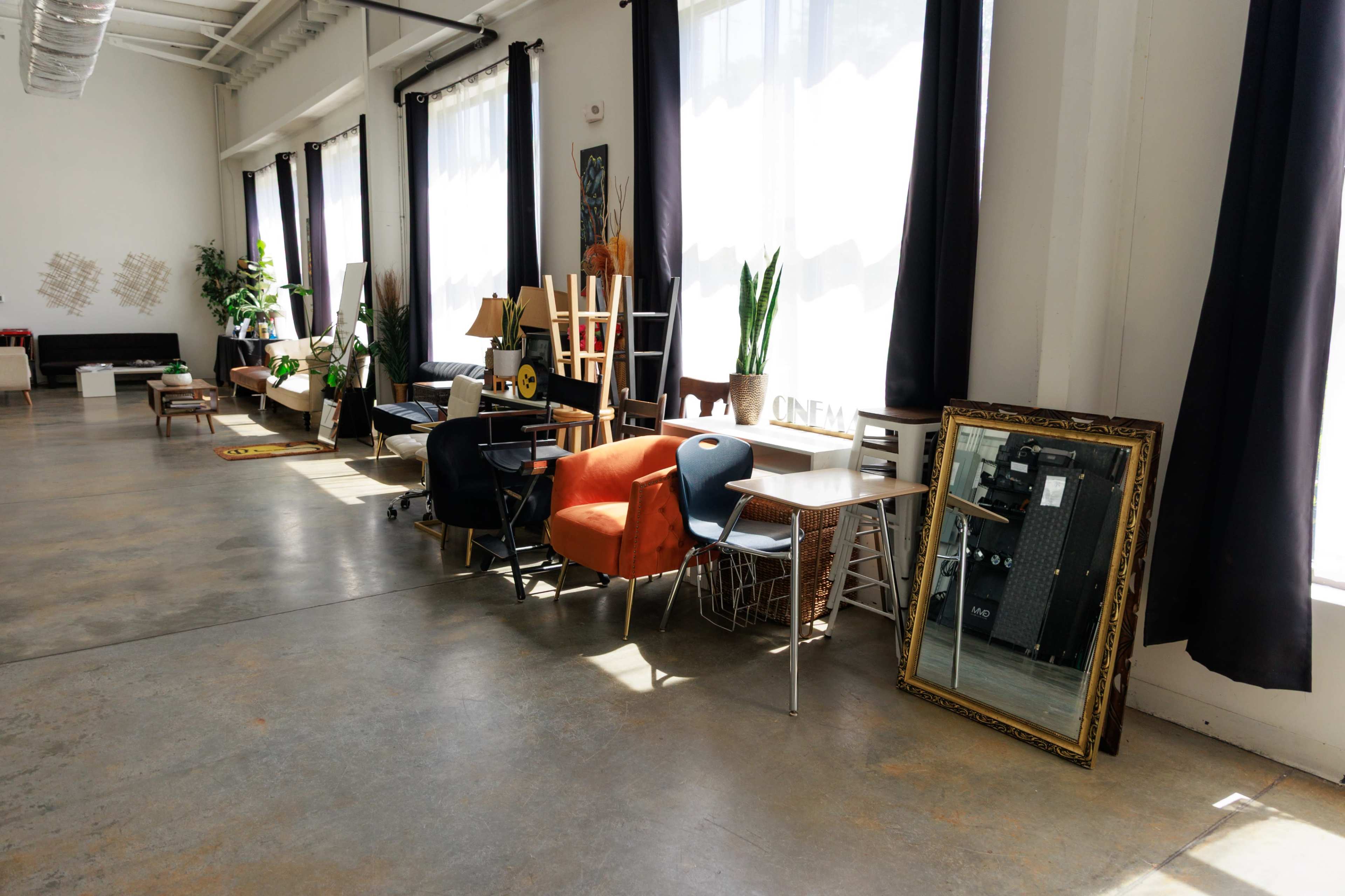 The image shows a sunlit indoor space with various pieces of furniture, including chairs, a table, and a large mirror, arranged along a polished concrete floor.