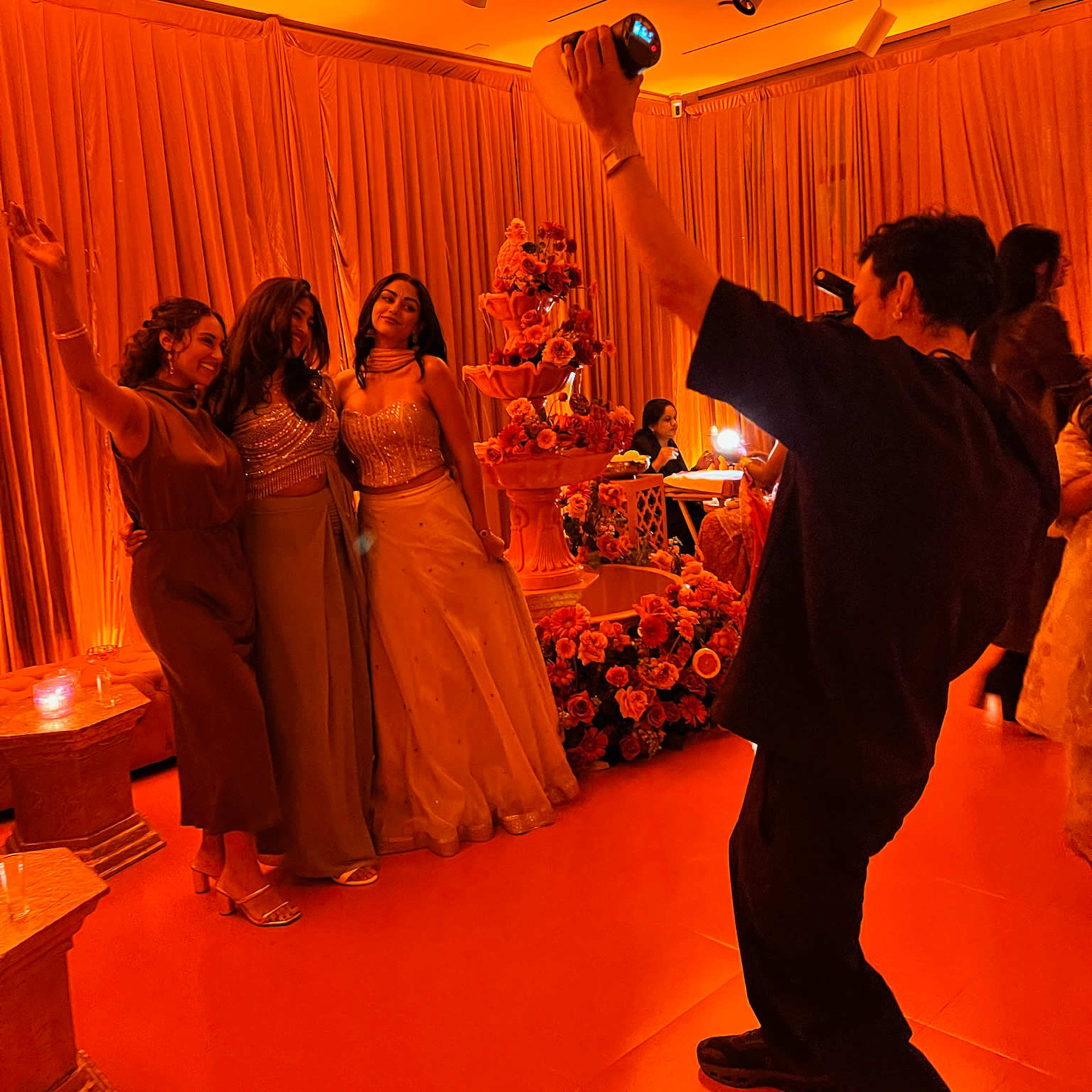 Three women pose for a photo while a man takes their picture in a decorated indoor setting with orange lighting and floral arrangements.