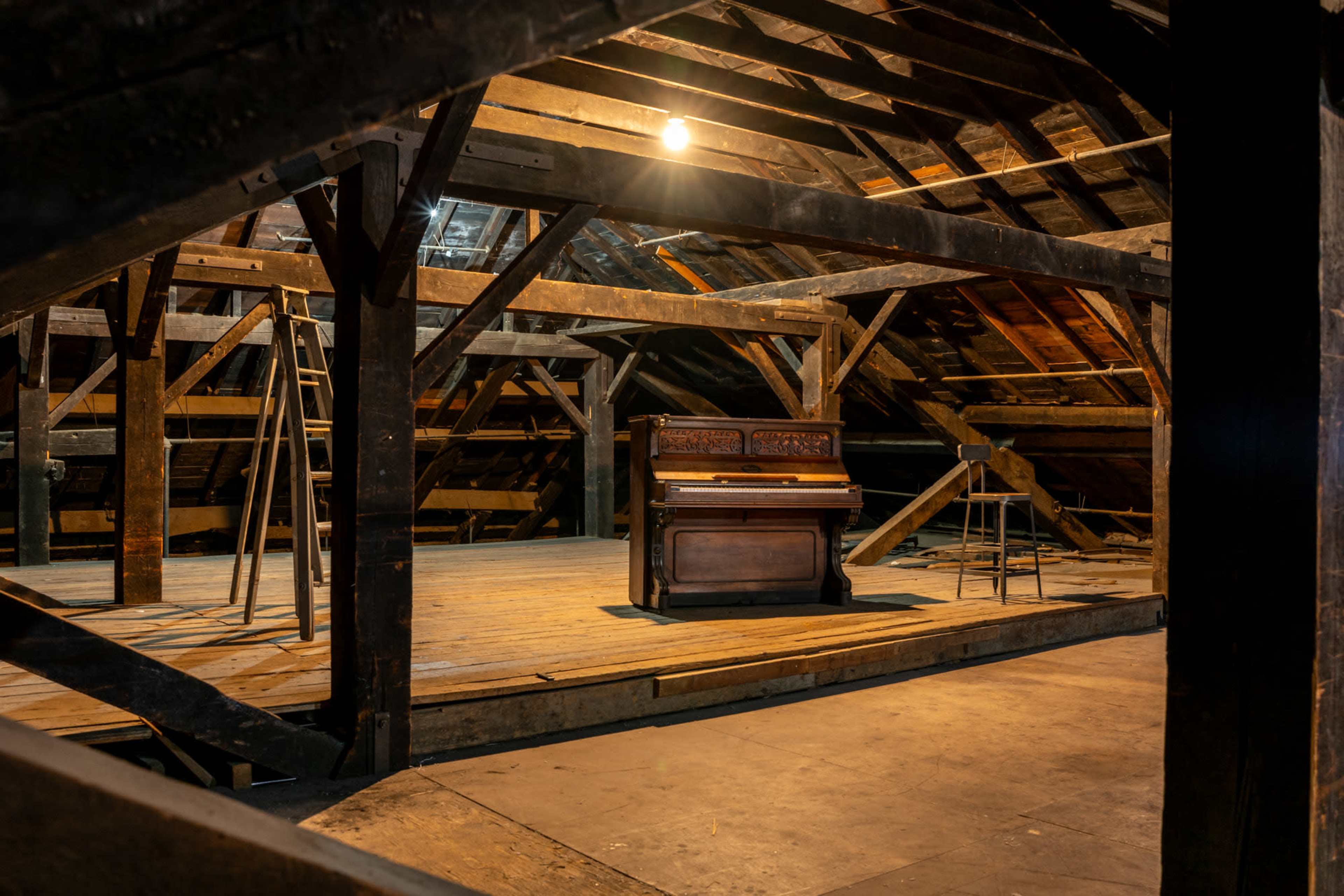 An old piano and a stool are positioned in a dimly lit attic with exposed wooden beams and a wooden floor.